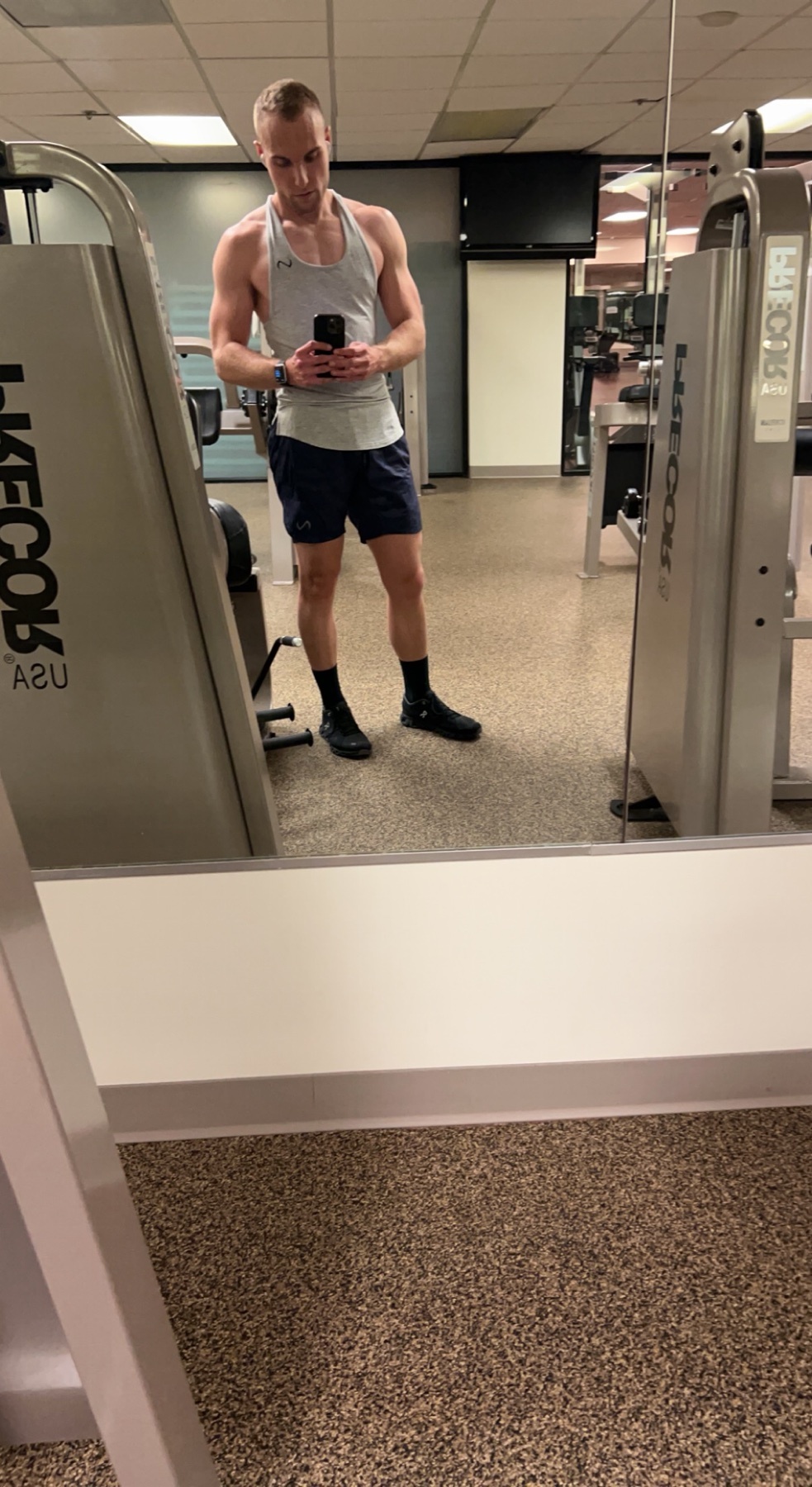 A man takes a mirror selfie in a gym, showcasing his physique. He's wearing a tank top and shorts, posing in front of gym equipment.