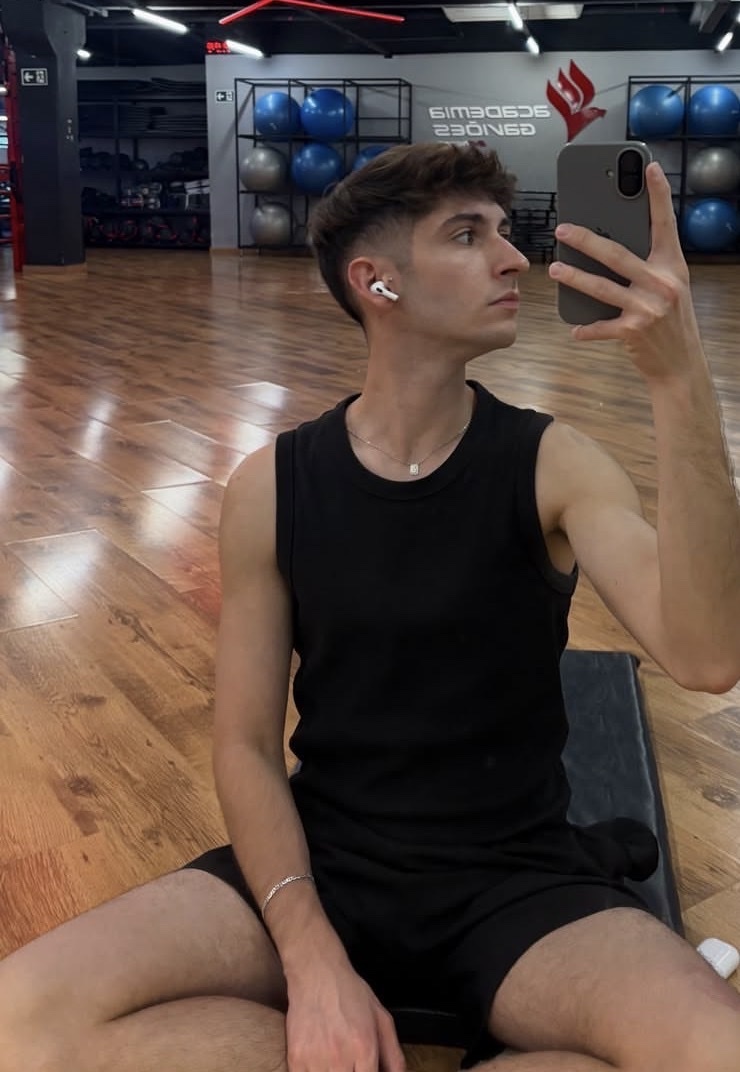 A young man takes a selfie in a gym, wearing a black tank top and shorts. He is sitting on the floor with earbuds in his ears.