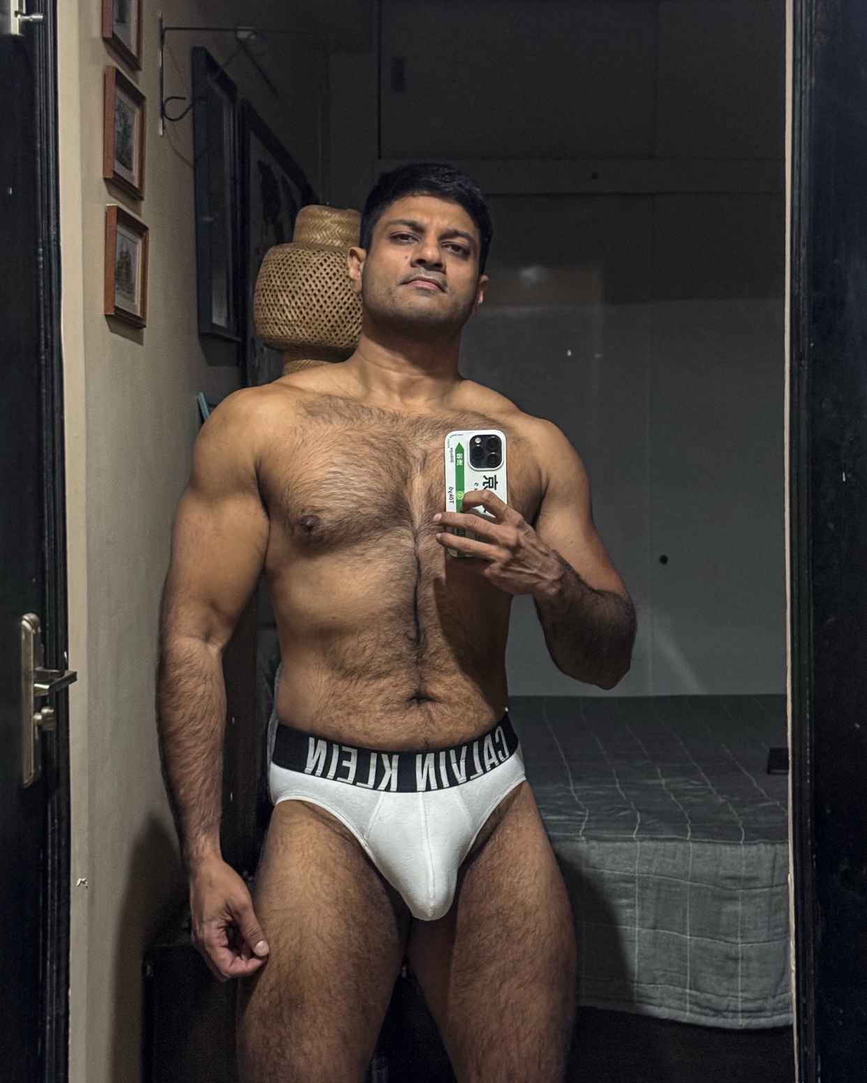 A man poses shirtless in front of a mirror, wearing white underwear. The photo highlights his muscular physique.