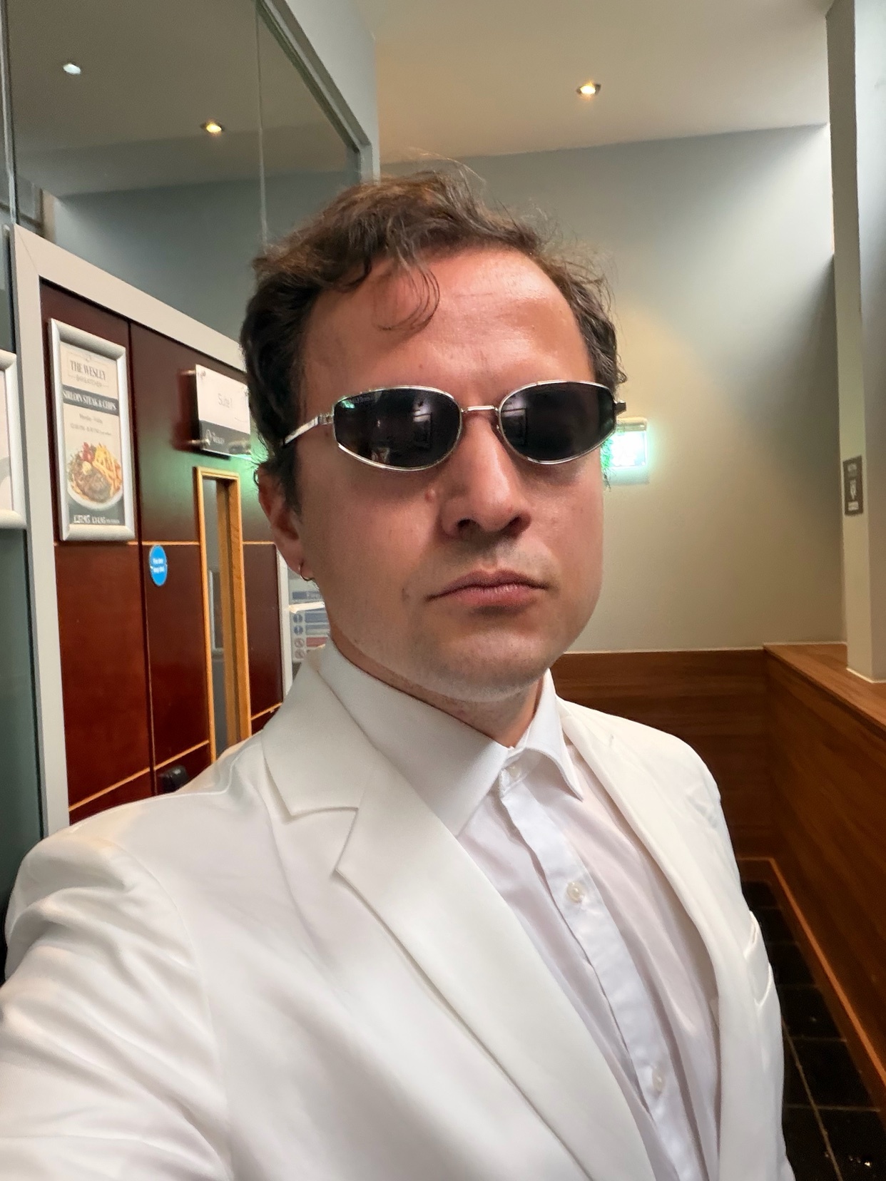 A man in a white suit and sunglasses takes a selfie indoors.