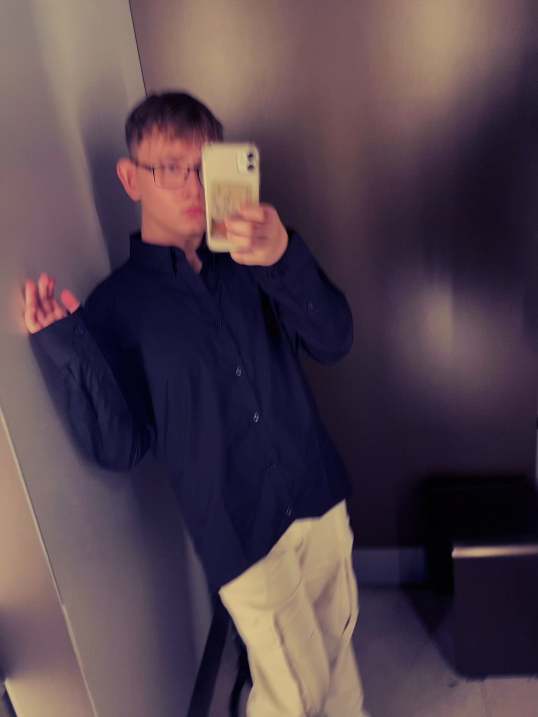 A young man takes a selfie in a mirror, wearing glasses and a button-down shirt. The photo has a slightly blurred effect.