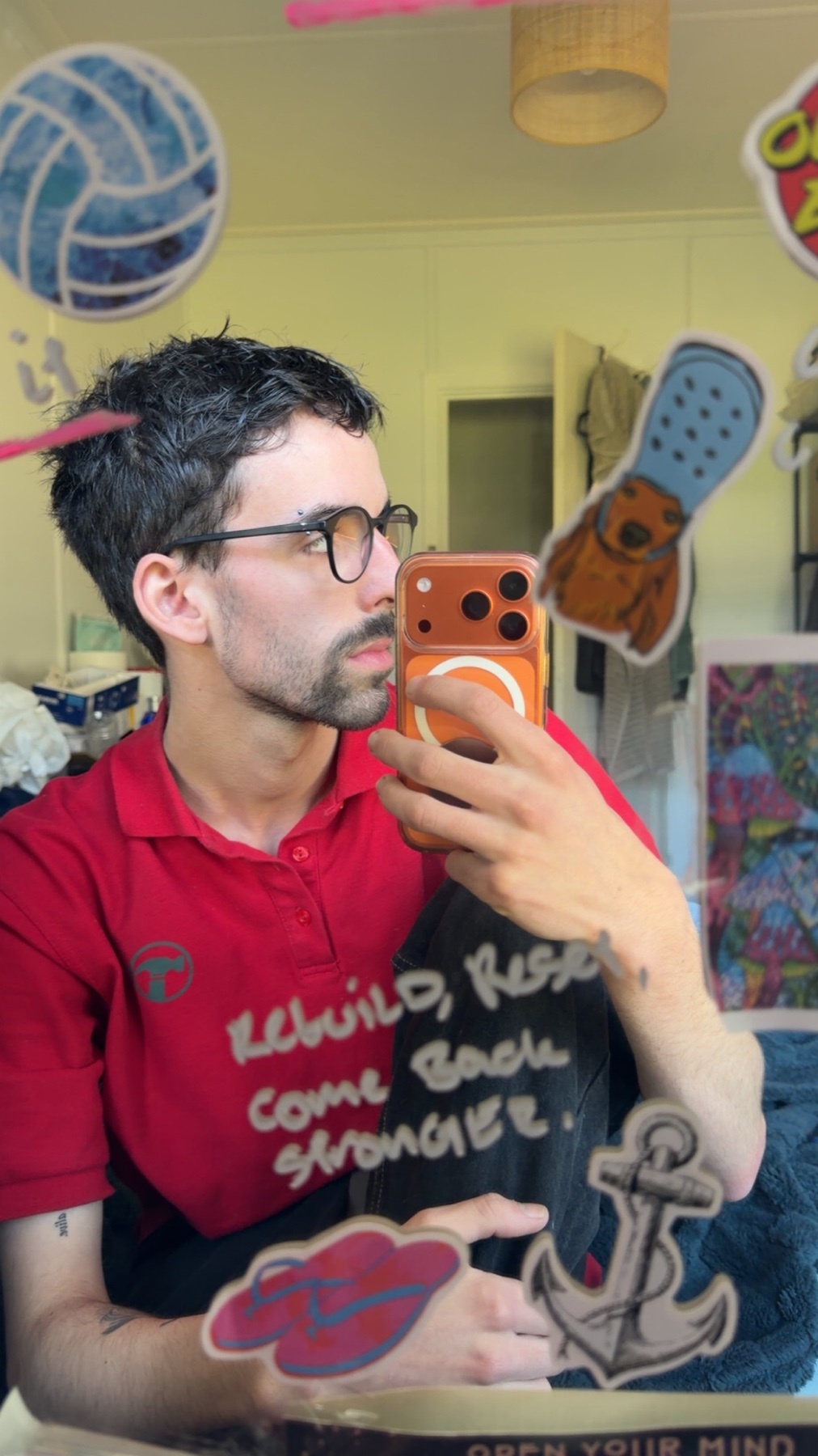 A man with glasses and stubble takes a mirror selfie. He is wearing a red shirt with an inspirational quote written on it in white marker. The mirror is decorated with various stickers.