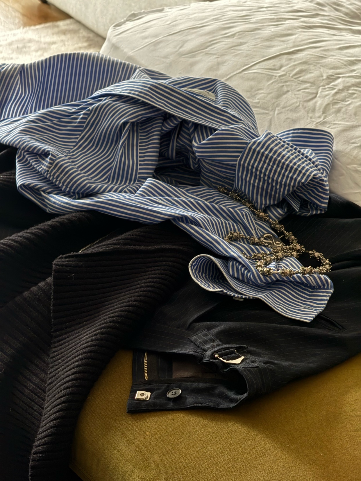 A close-up shot of a pile of men's clothing, including a striped shirt, black corduroy jacket, and a necklace. The items are arranged on a bed.