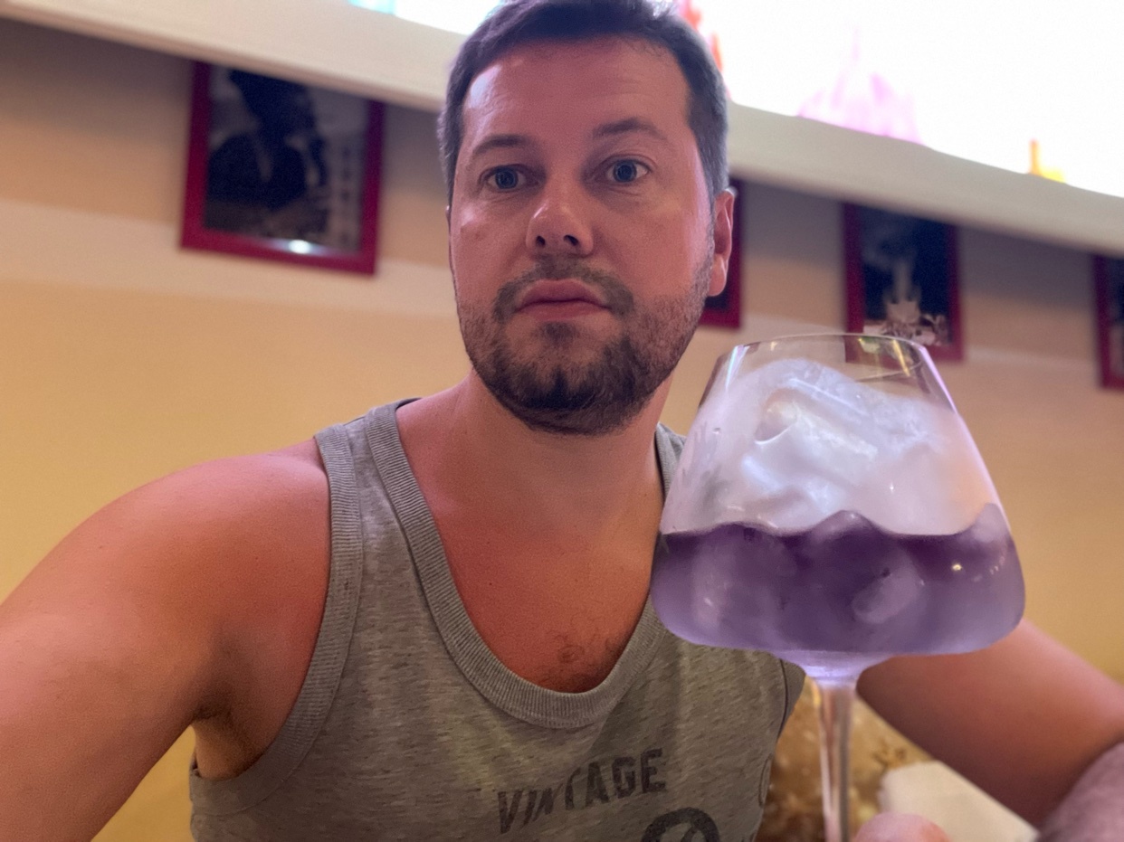 A man holds a purple cocktail in a glass. He is shirtless and looking at the camera.