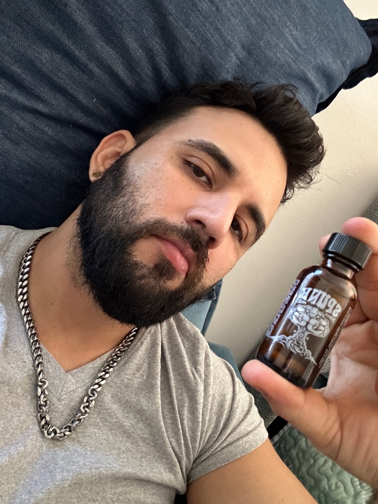 A man holds up a small bottle, looking directly at the camera. He has a beard and is wearing a grey shirt and a chain.
