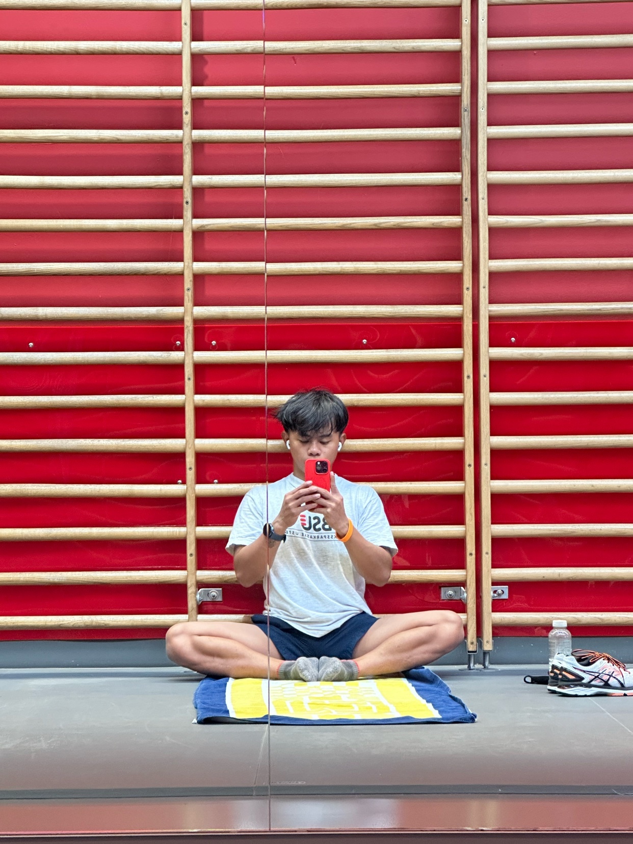 A man is sitting cross-legged on a mat, taking a mirror selfie. He is in a gym setting, with a red wall in the background.