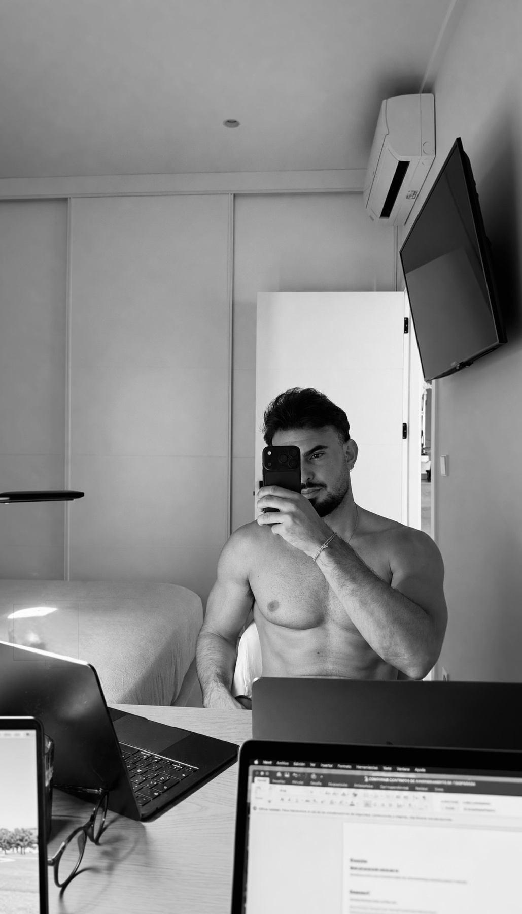 A shirtless man with a beard and stubble takes a selfie in black and white while sitting at a desk with laptops and glasses.