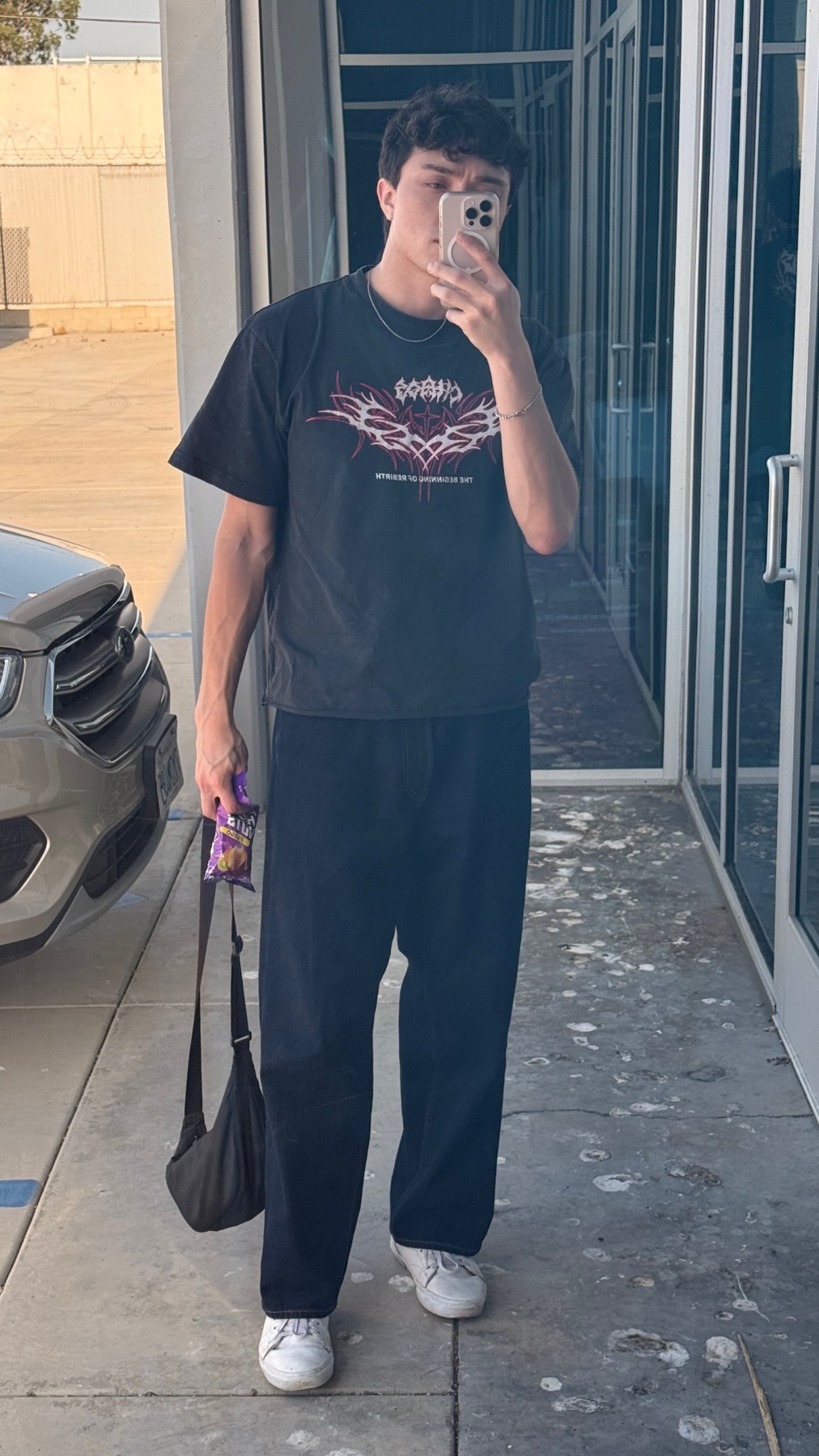 A man takes a selfie in a black graphic t-shirt and dark wash jeans. He is holding a small bag and a snack.