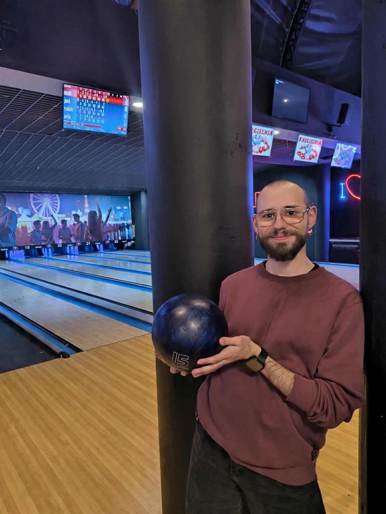 A man poses with a bowling ball in a bowling alley. He is smiling and looking at the camera.