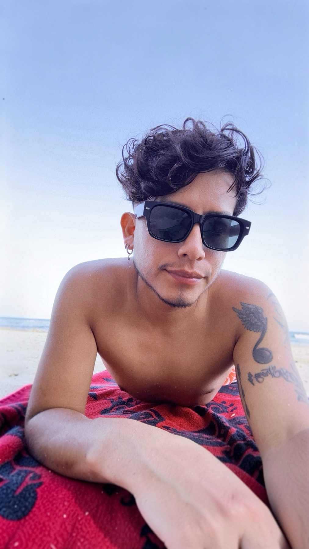 A man with curly dark hair and sunglasses lies on a red blanket on the beach. He has tattoos on his arm and stubble on his face.