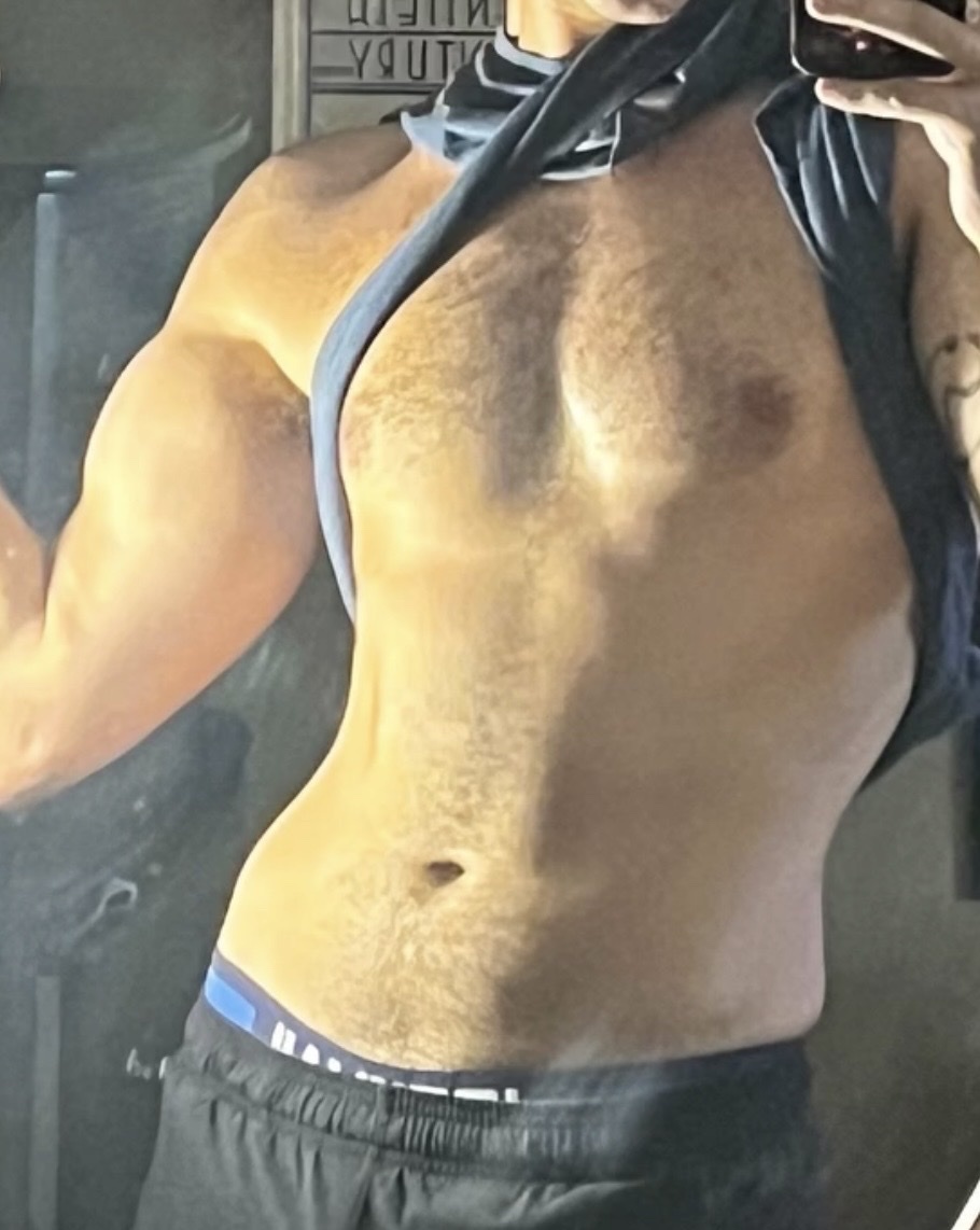 A man shows off his muscular physique, flexing his bicep and revealing his abs. He is wearing black athletic shorts and has a tattoo on his arm.
