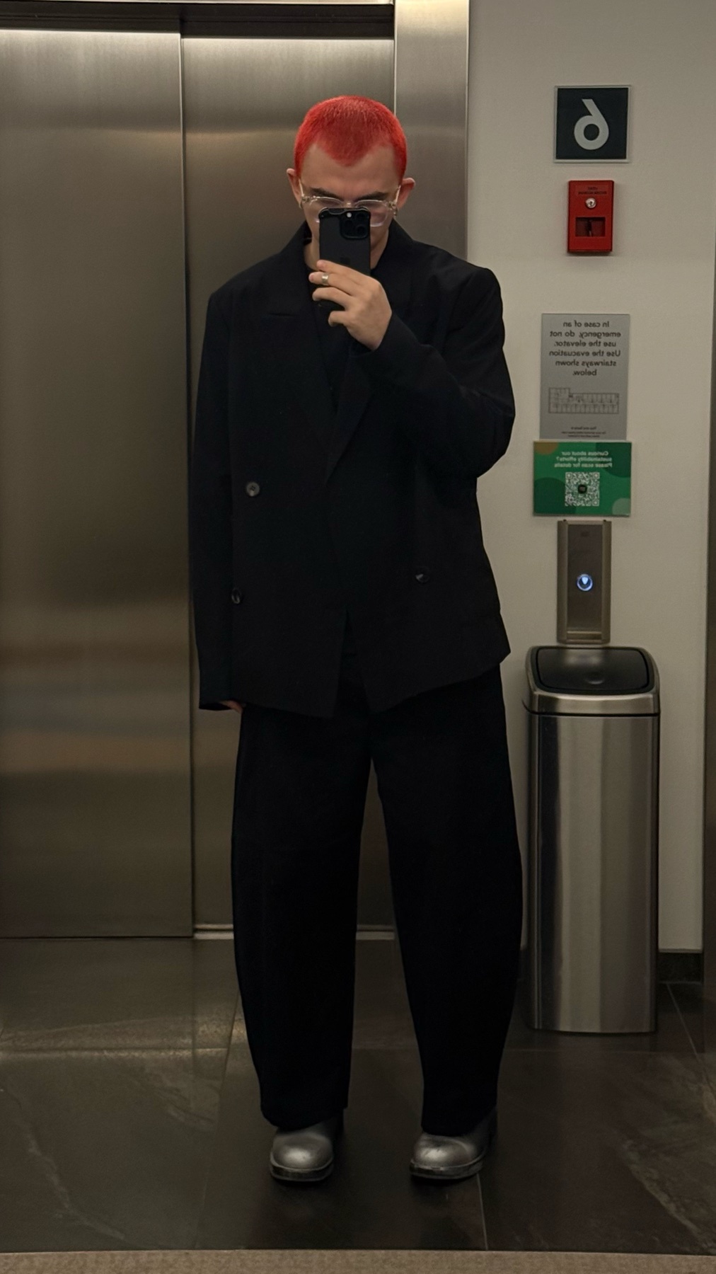 A man with bright red hair and glasses takes a selfie in an elevator while wearing a black suit and silver shoes.