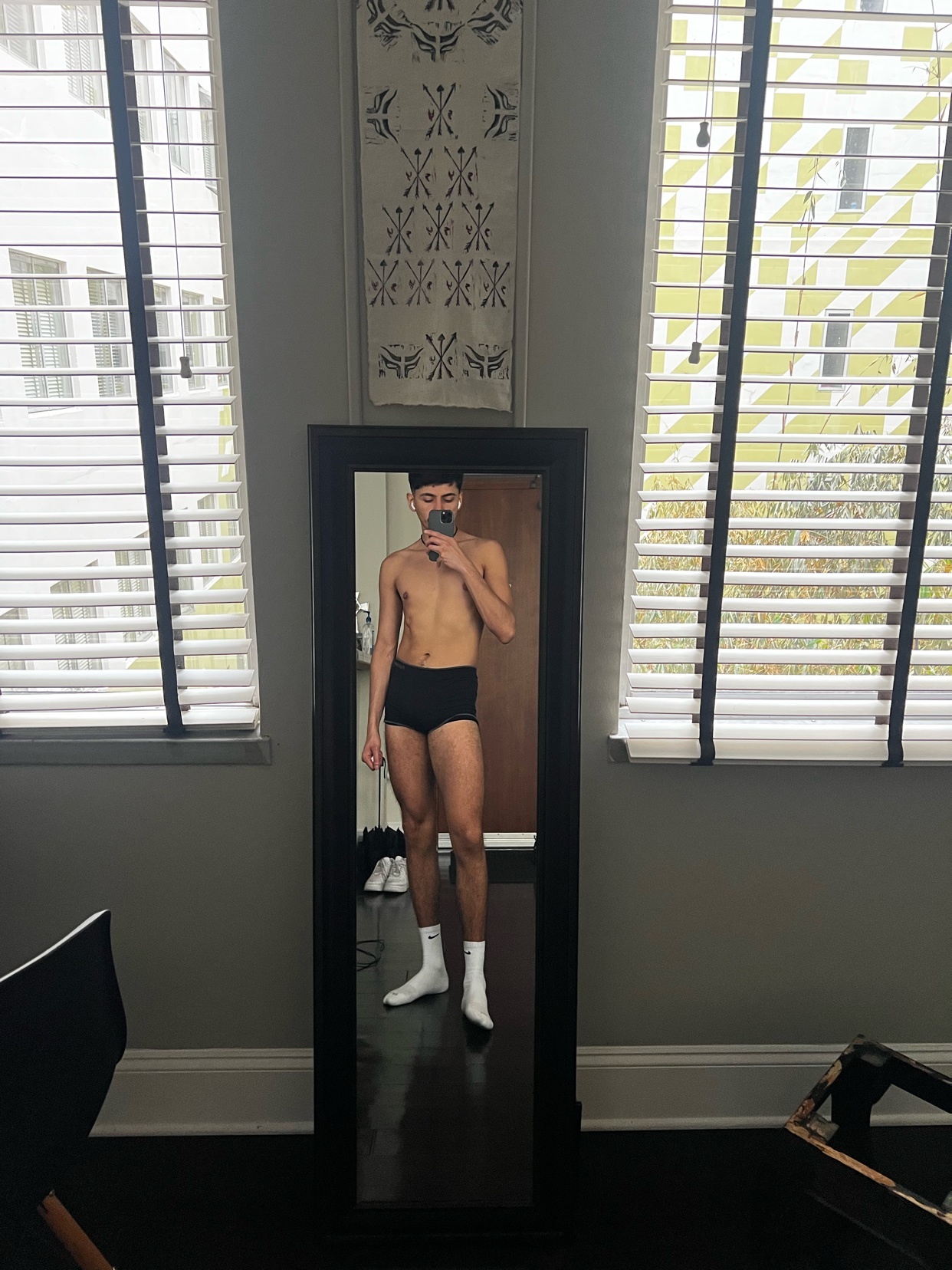 A man poses shirtless in front of a mirror, wearing black underwear and white socks. The photo is taken indoors, with a window and blinds in the background.