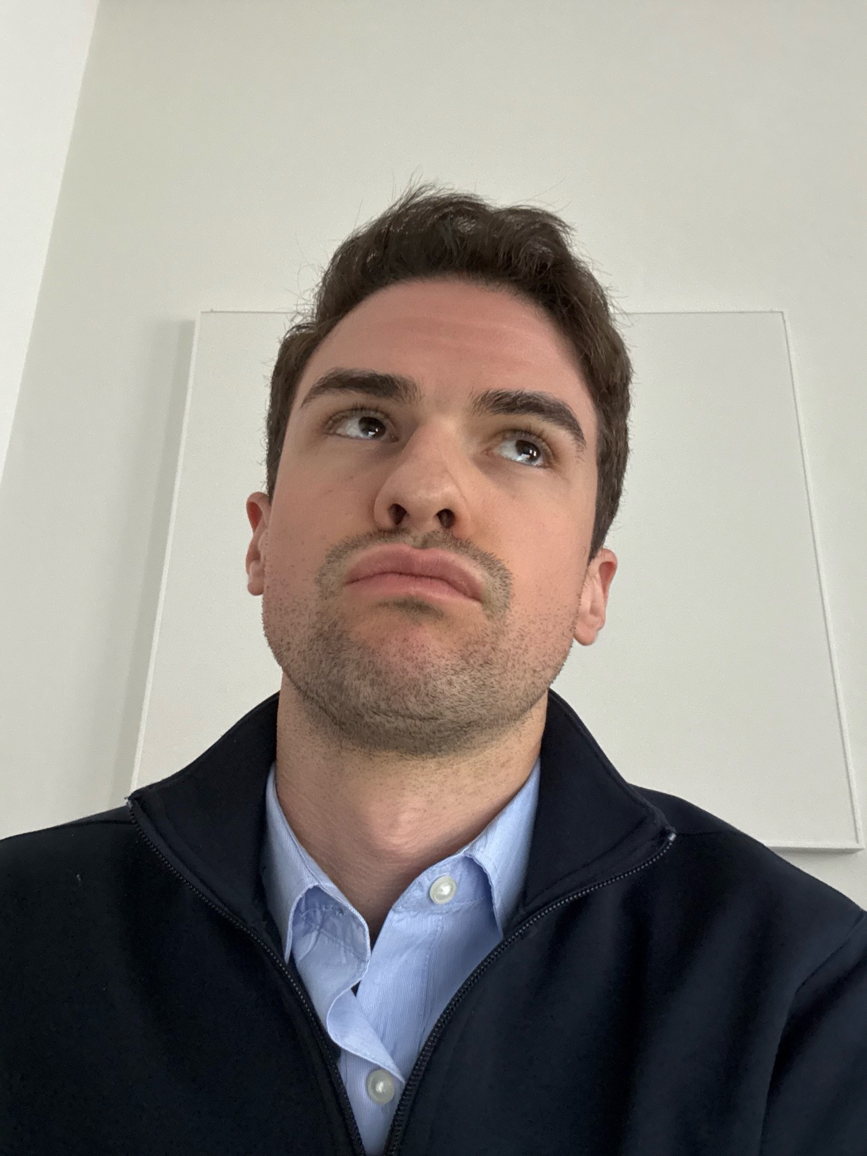 A man looks up with a serious expression. He is wearing a blue shirt and a dark jacket.