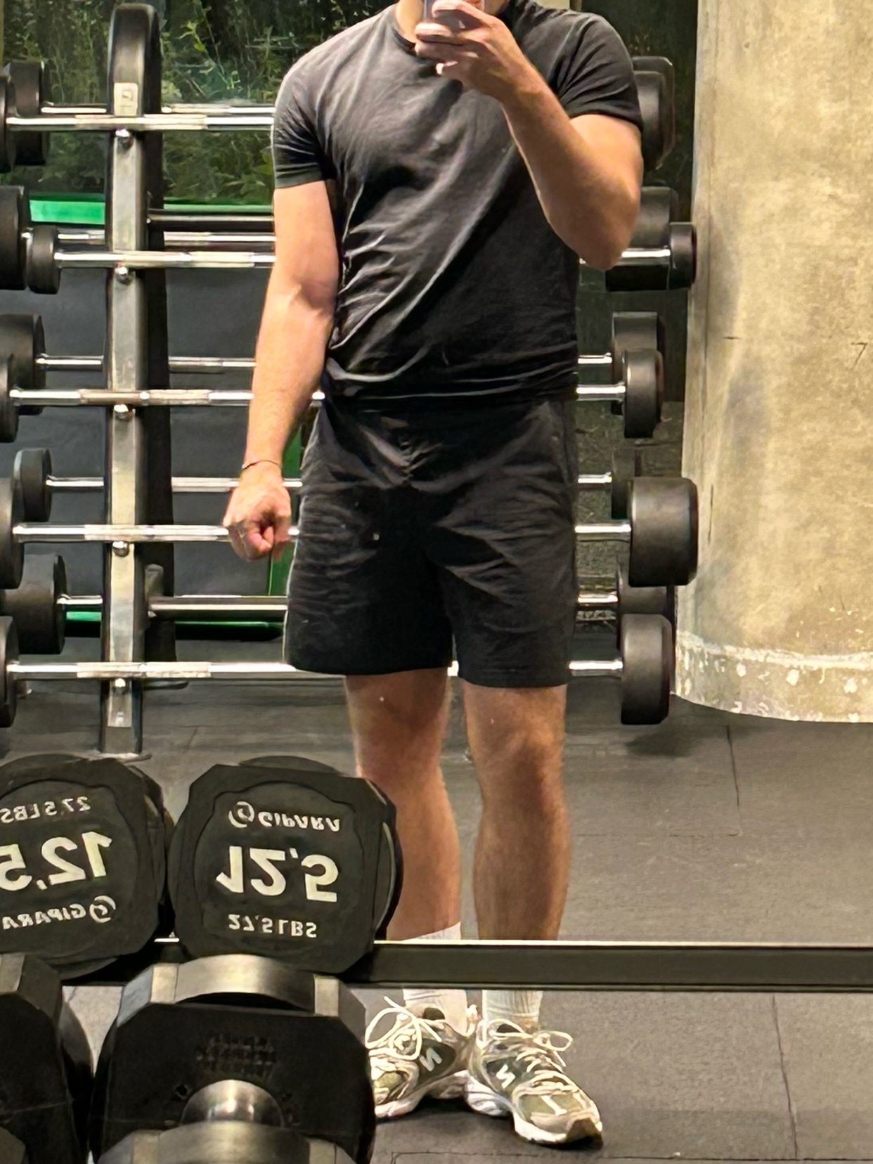 A man poses in a gym mirror, showcasing his physique. He is wearing a black t-shirt and shorts.