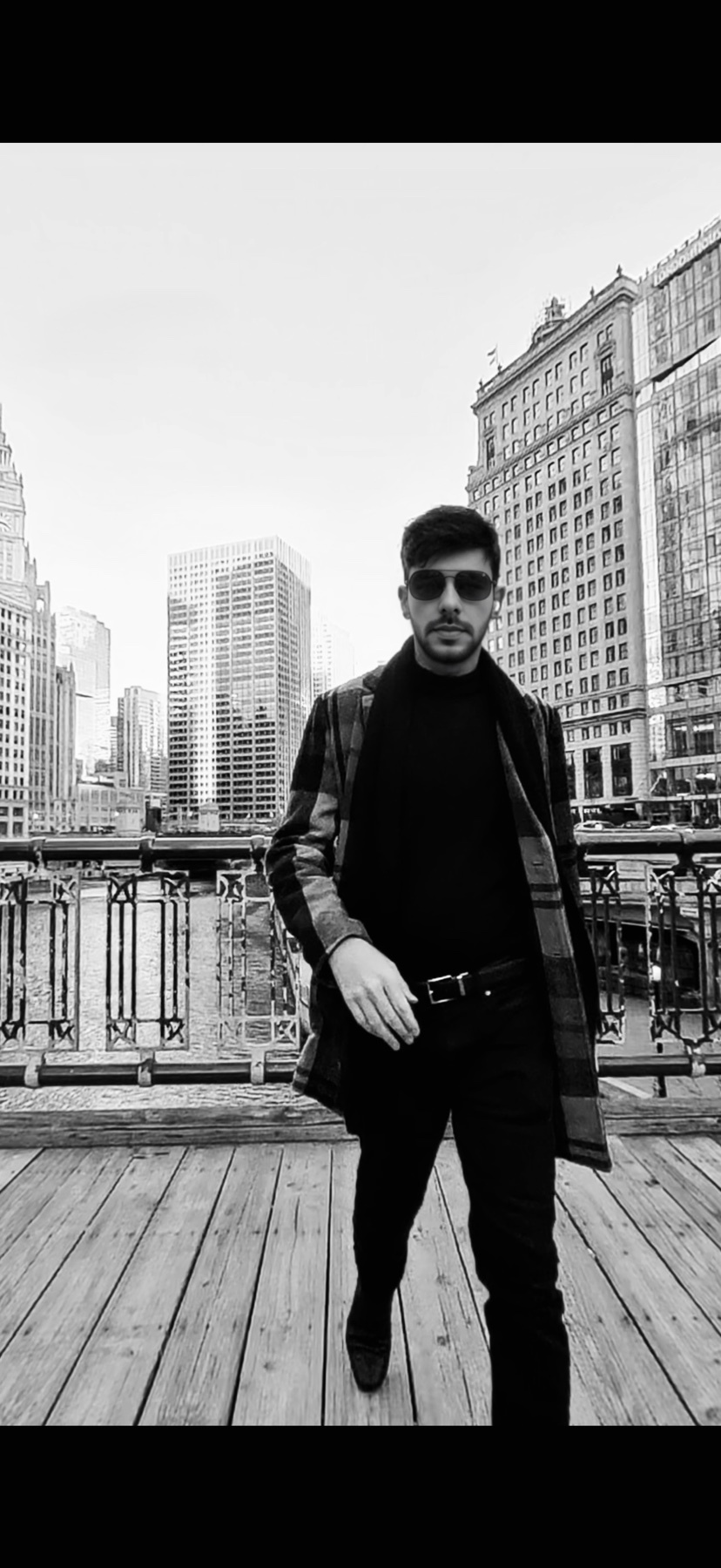 A man walks on a wooden boardwalk in a city, wearing sunglasses and a stylish coat over a turtleneck and dark pants. The black and white image captures a cityscape with tall buildings in the background.
