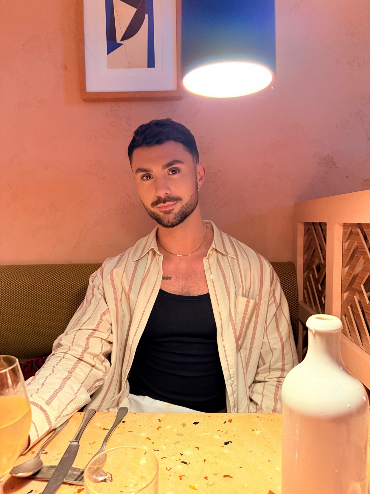 A man poses for a photo at a restaurant. He is wearing a striped shirt over a black tank top.