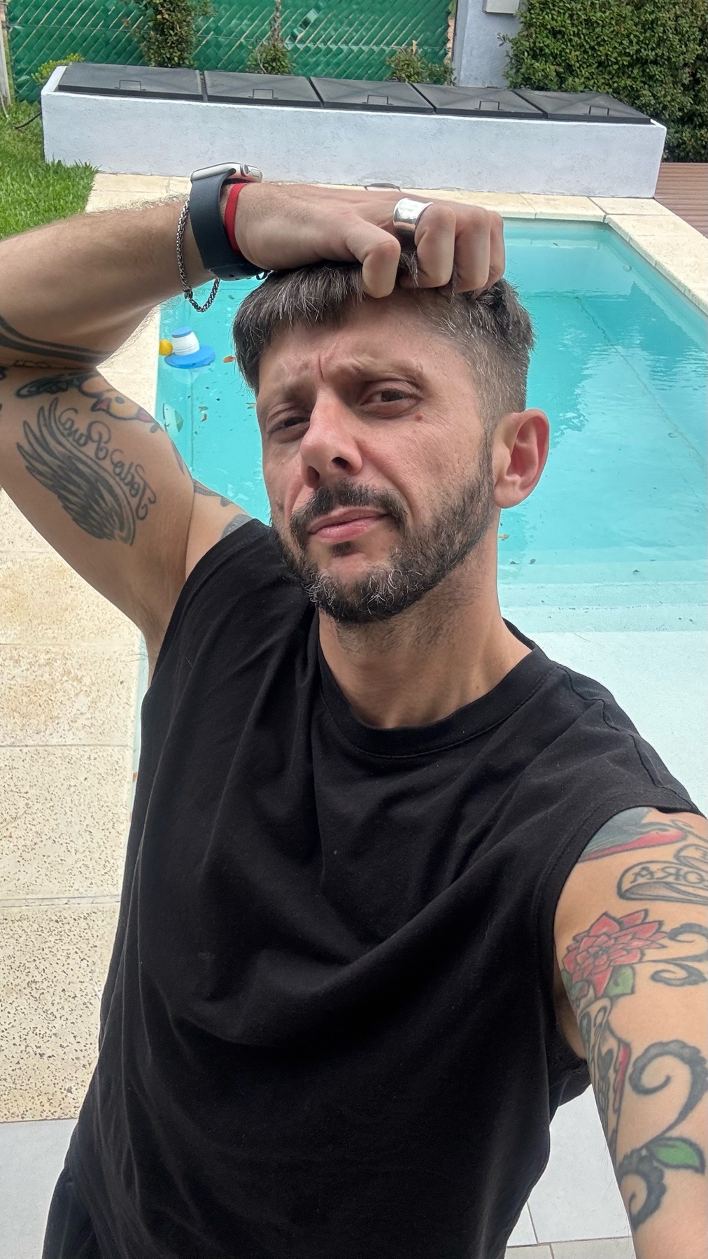 A man poses for a selfie by a pool, showing off his tattoos and physique. He is shirtless, with a sleeveless shirt, and has a serious expression.