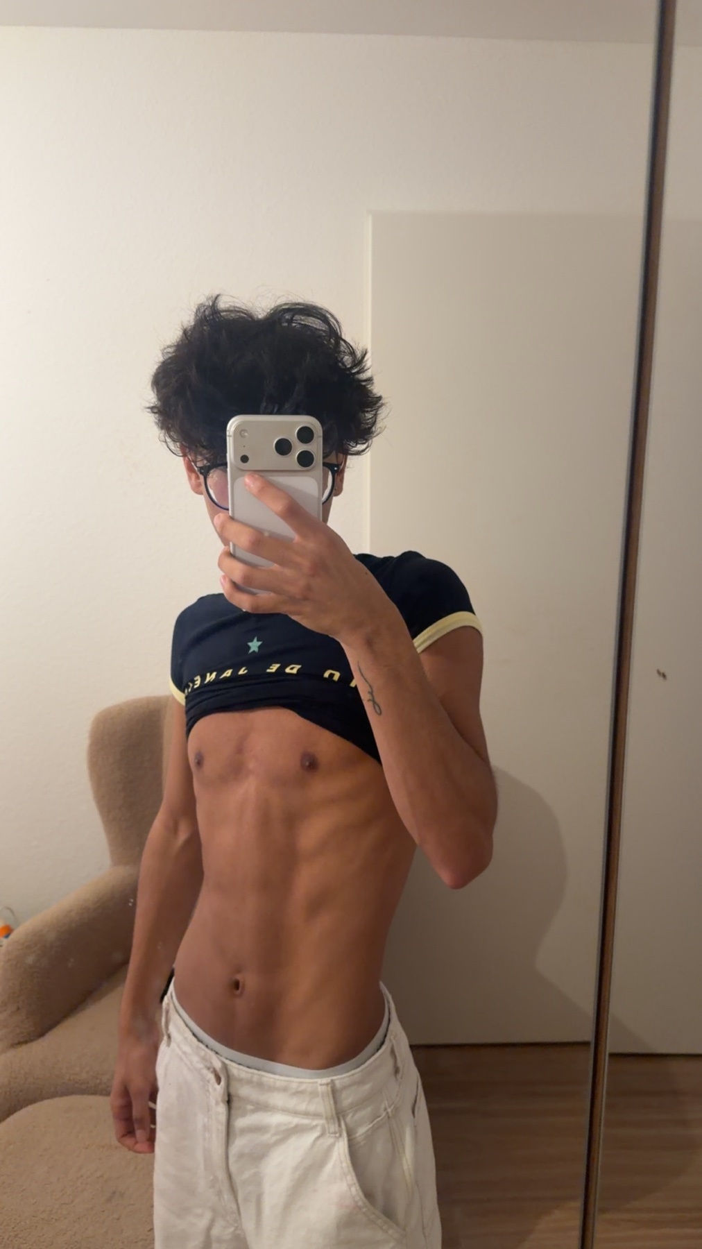 A person takes a mirror selfie in a casual outfit, showing off their abs. The person is wearing white pants and a dark t-shirt that is pulled up to reveal their bare torso.