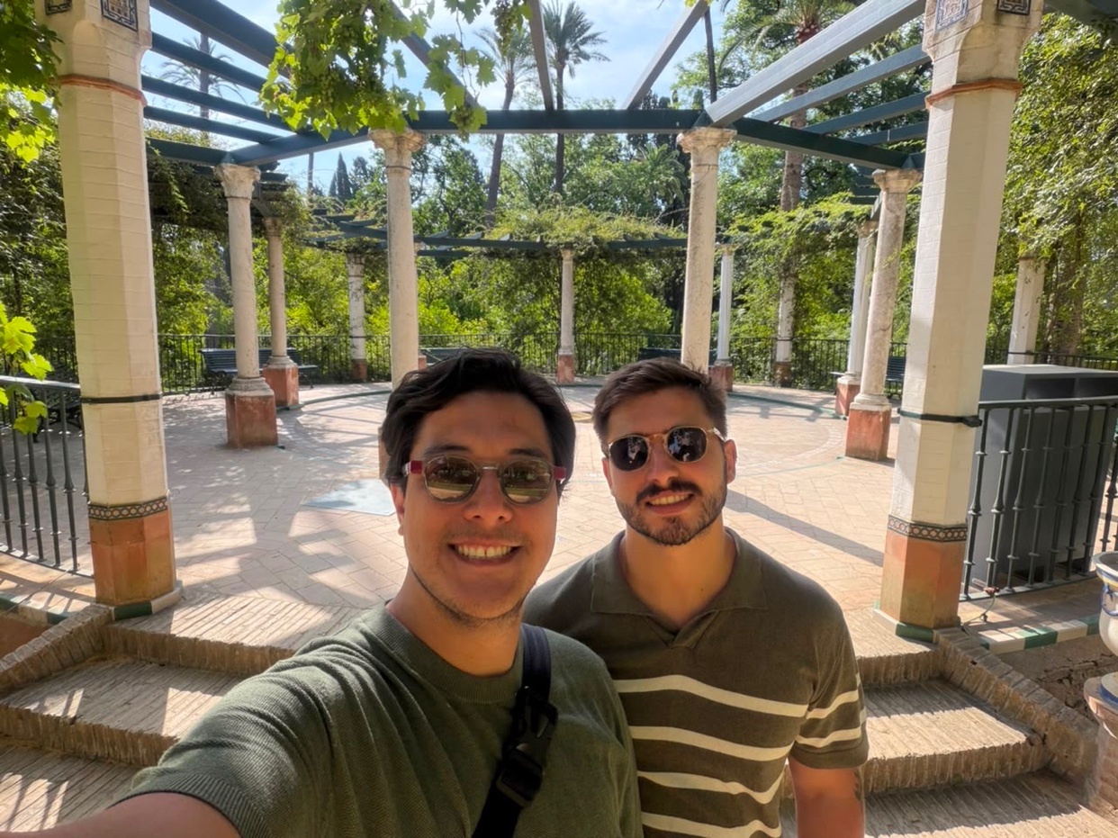A selfie of two men smiling in front of a beautiful outdoor pavilion with columns and lush greenery.
