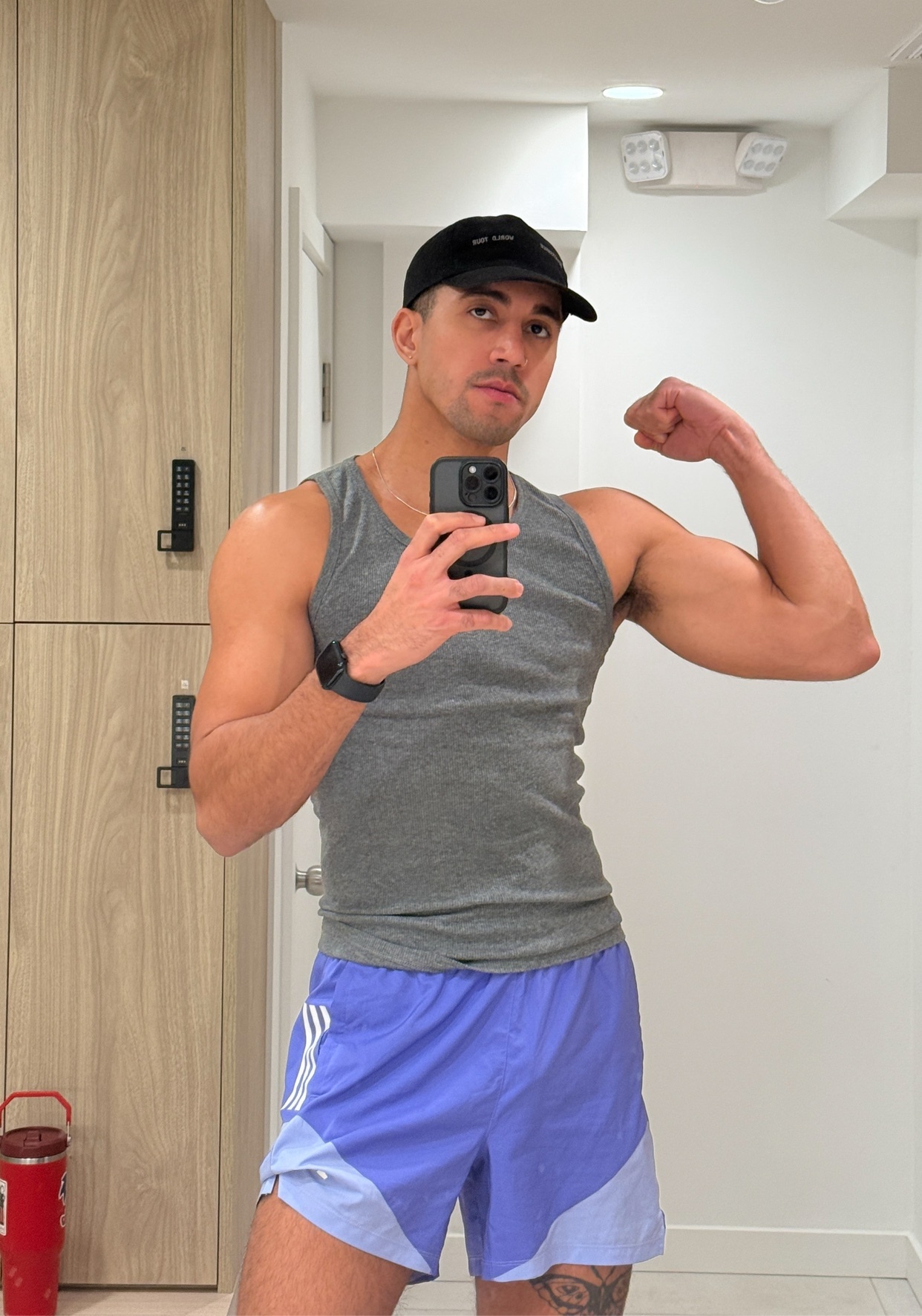 A man flexes his bicep in a mirror selfie. He is wearing a tank top and shorts.