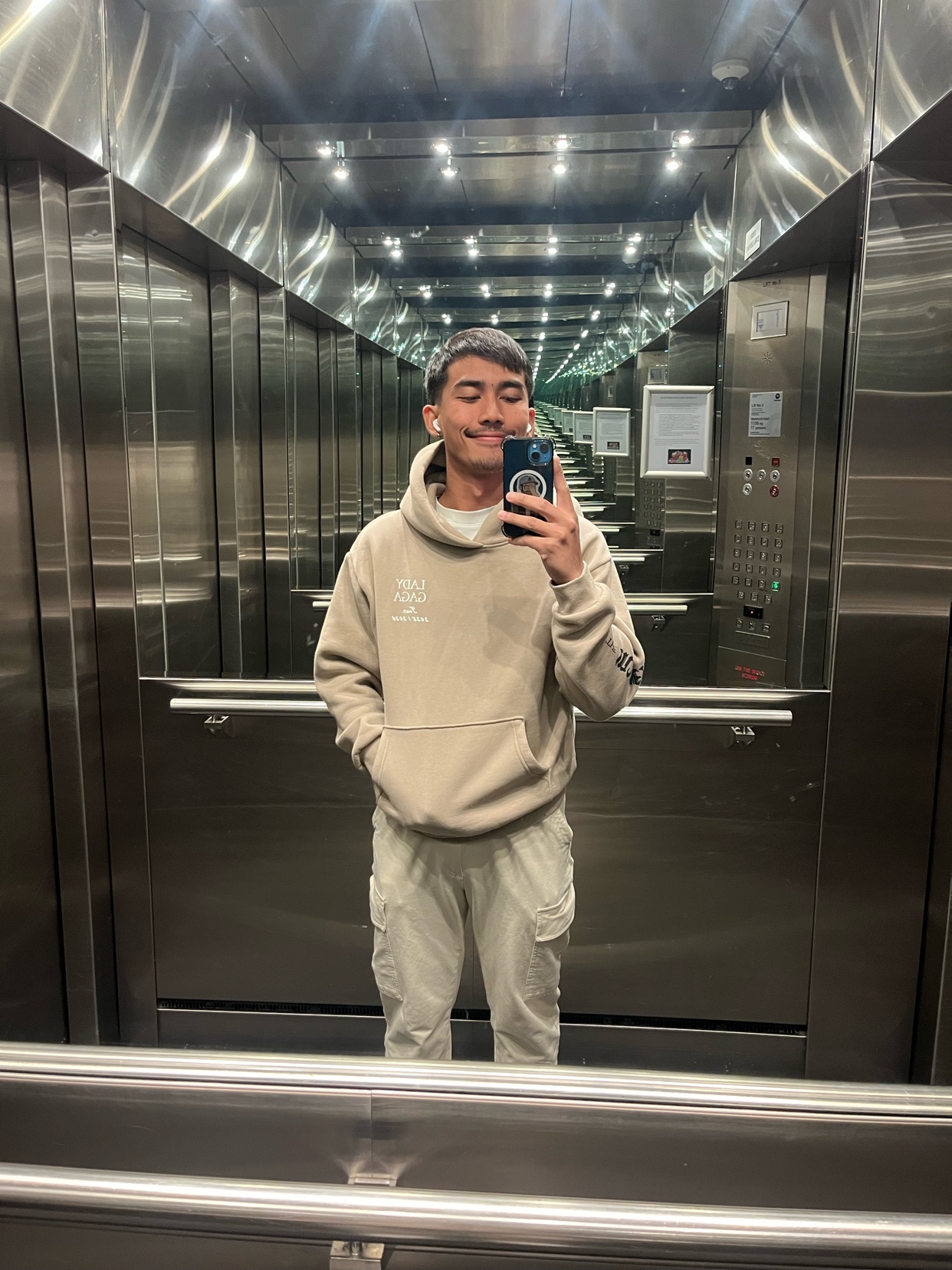 A man takes a selfie in an elevator. He is wearing a beige hoodie and matching pants.
