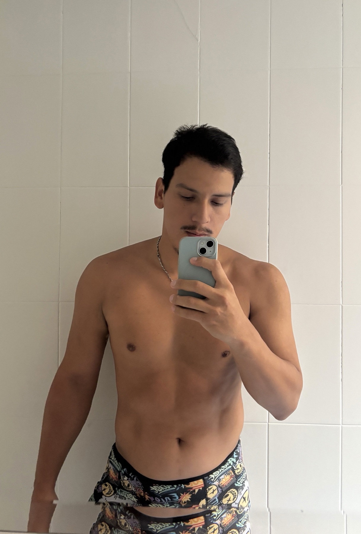 A man takes a mirror selfie in a bathroom, wearing patterned shorts and no shirt.