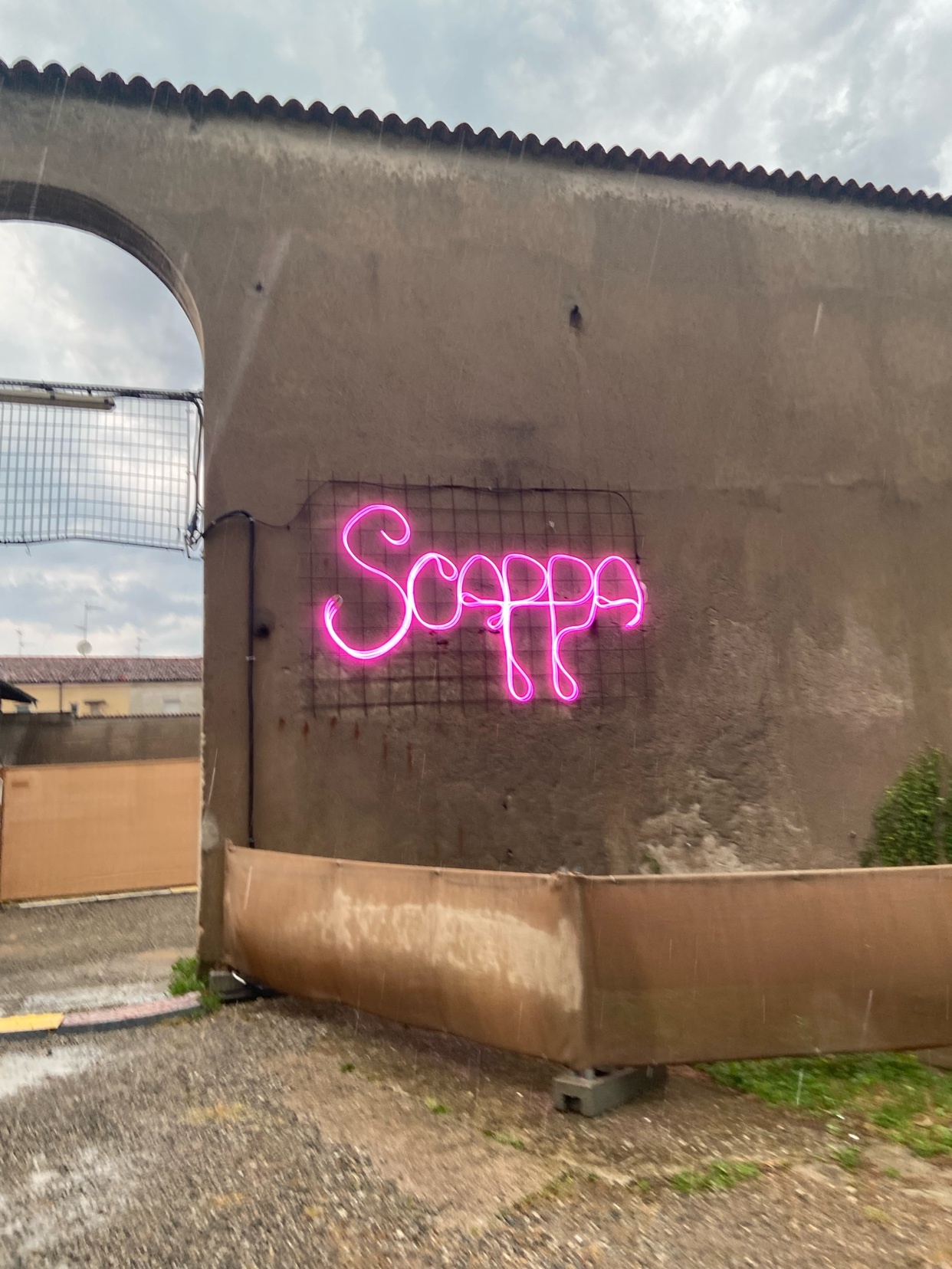 A bright pink neon sign spelling out "Scoppa" is mounted on a textured, brown wall under a cloudy, rainy sky. The sign is attached to a black wire grid, and the surrounding area appears to be an outdoor space with an archway visible to the left.