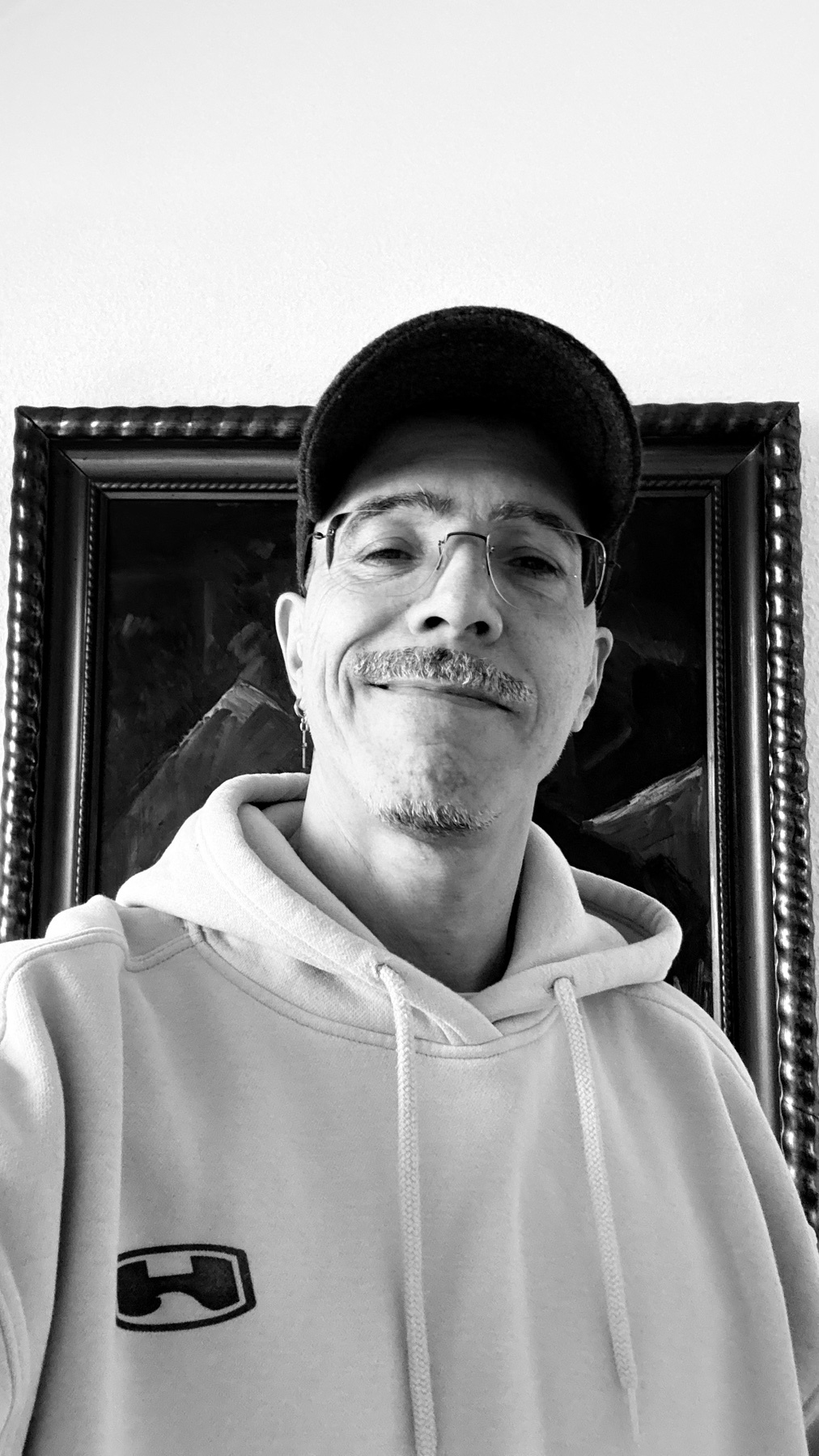 A black and white selfie of a man wearing glasses, a baseball cap, and a hoodie. He has a mustache and a goatee.