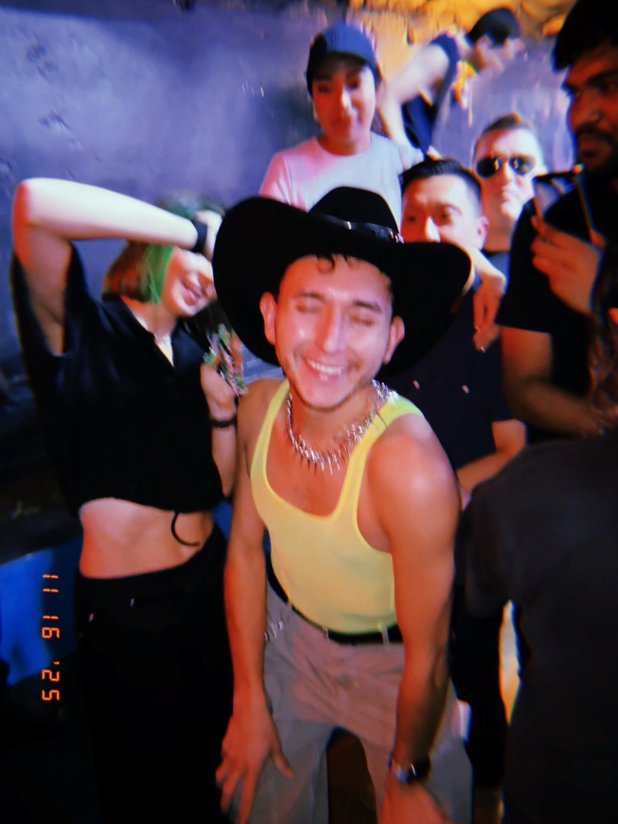 A group of friends pose for a photo at a party. A man in a cowboy hat and tank top smiles at the camera.