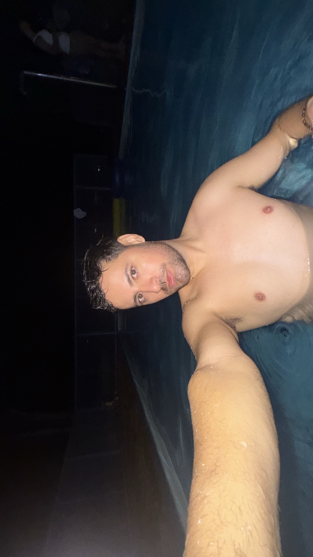 A man takes a selfie in a pool at night. He is shirtless and has dark hair and stubble.