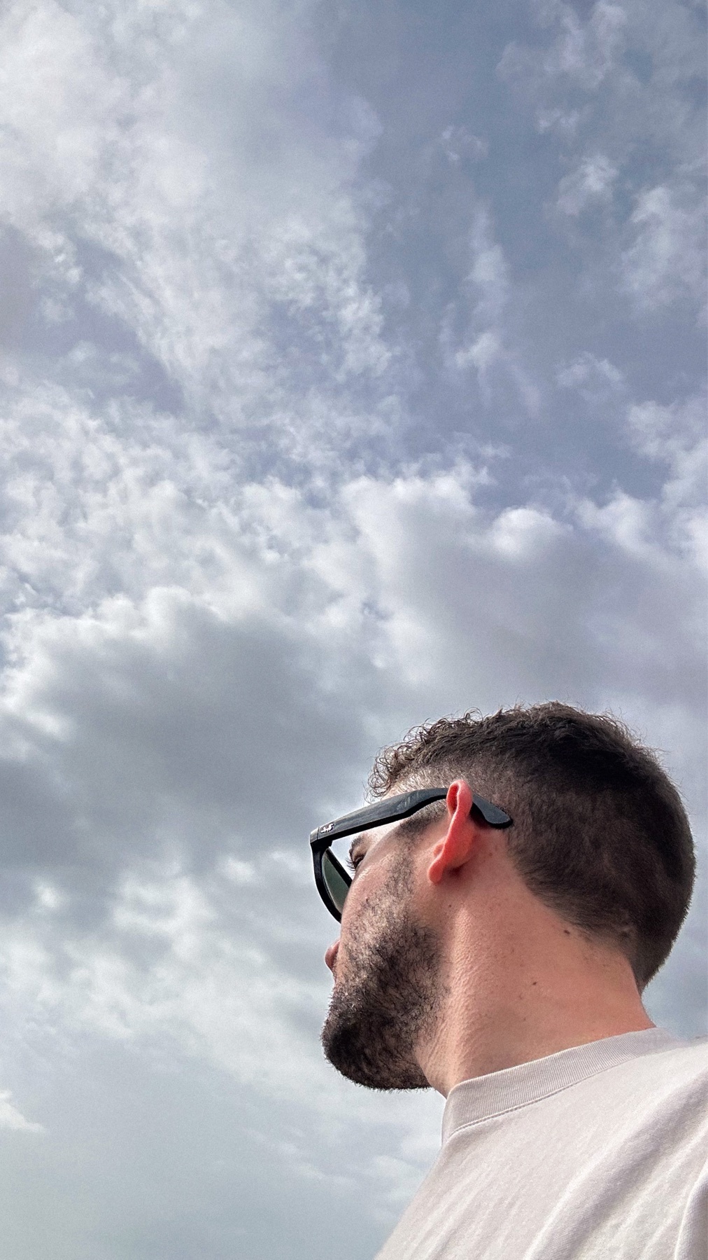 A man wearing sunglasses looks up at a cloudy sky. The photo is a portrait of the man from the side.