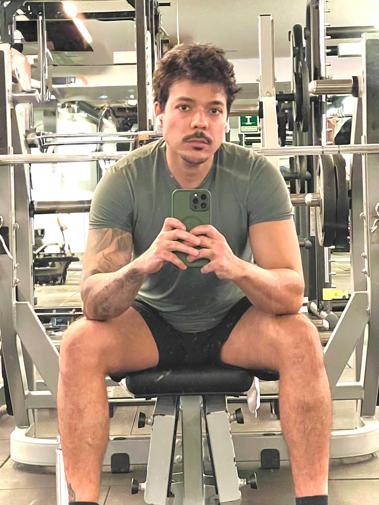A man with a mustache and tattoos sits on a weight bench in a gym, holding his phone to take a selfie.