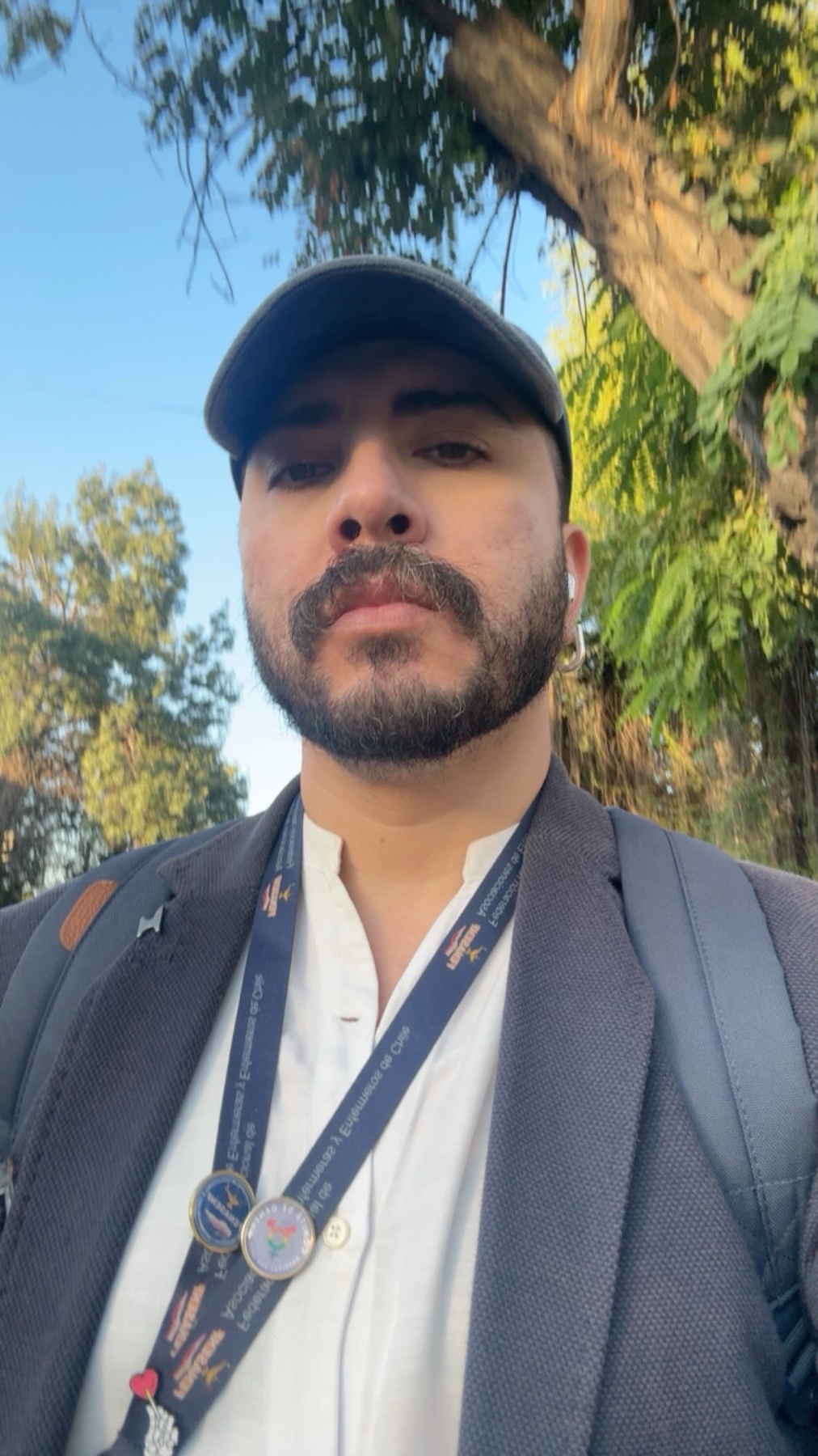 A man wearing a baseball cap and a blazer over a white shirt poses for a selfie outdoors.