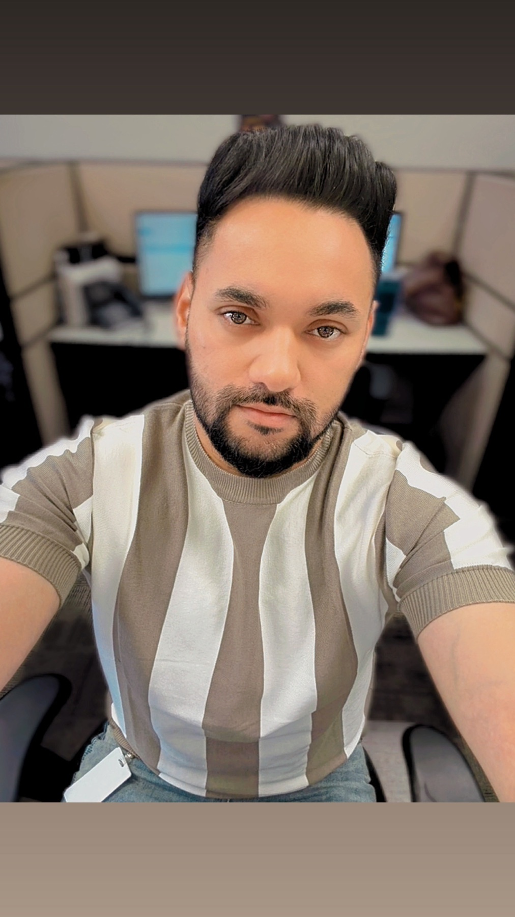 A man with a beard and stubble takes a selfie in an office cubicle. He is wearing a vertically striped shirt.