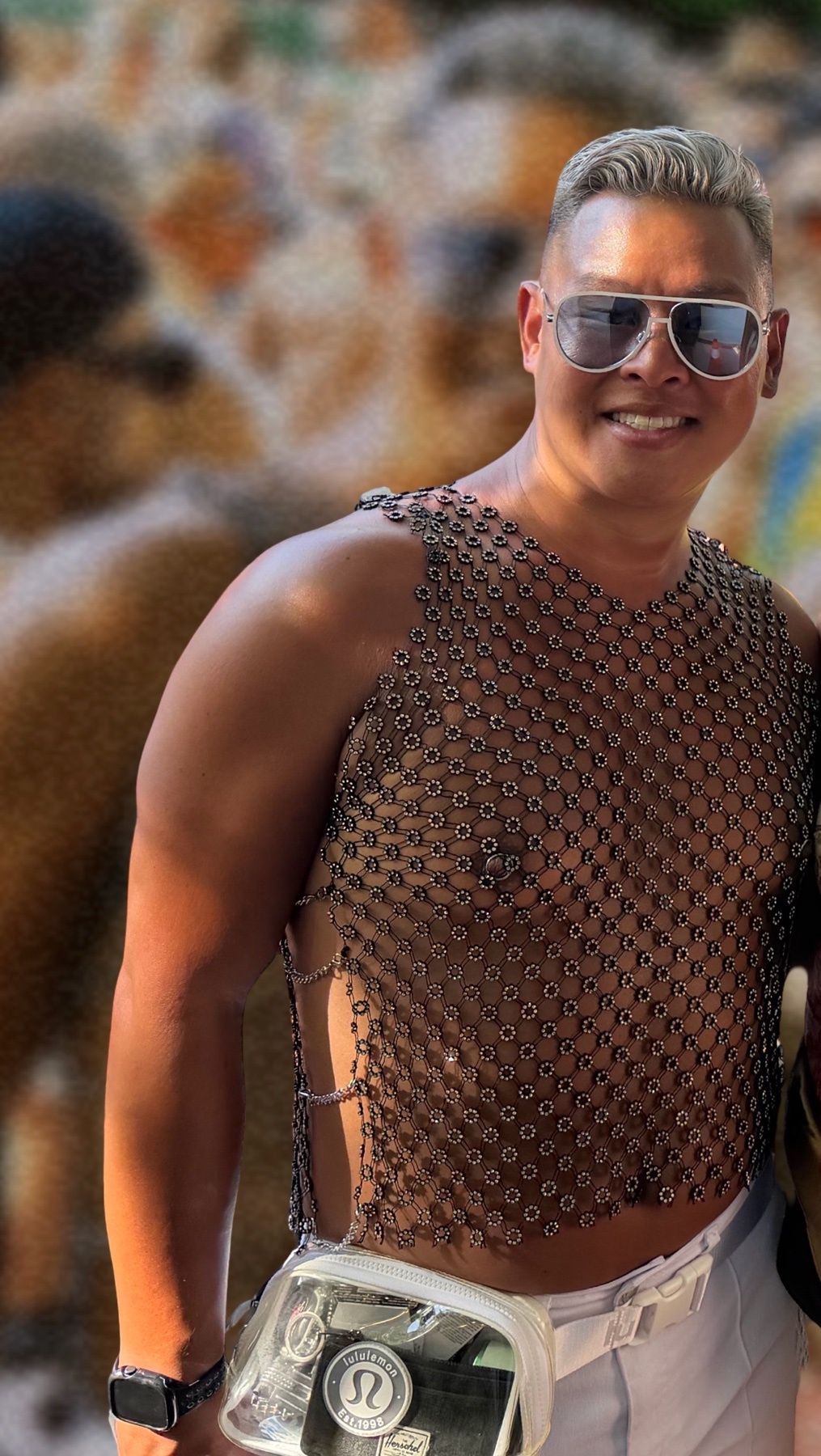 A man smiles at the camera wearing a chainmail top and sunglasses. The background is blurred, suggesting a crowded event.