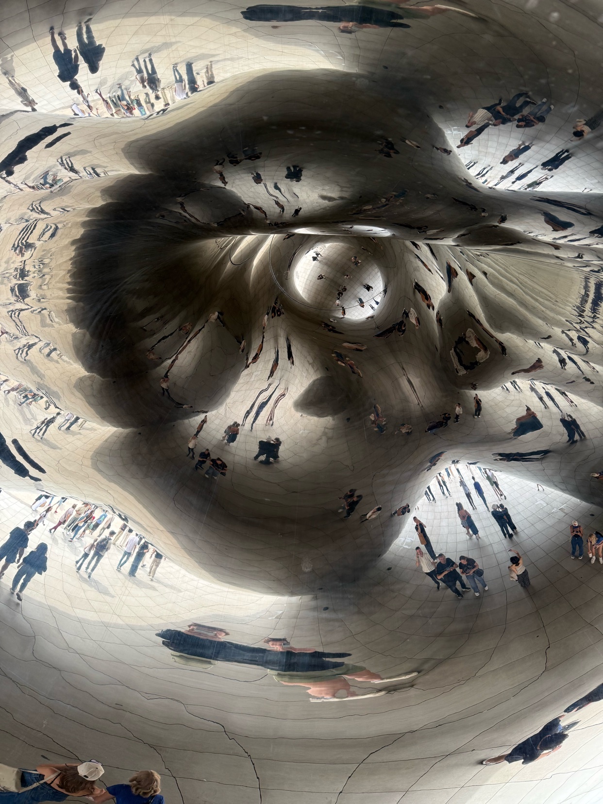 A surreal, distorted reflection of people and the surrounding environment is captured on a large, metallic sculpture. The artwork creates a warped and abstract visual experience, reflecting the visitors in a unique way.