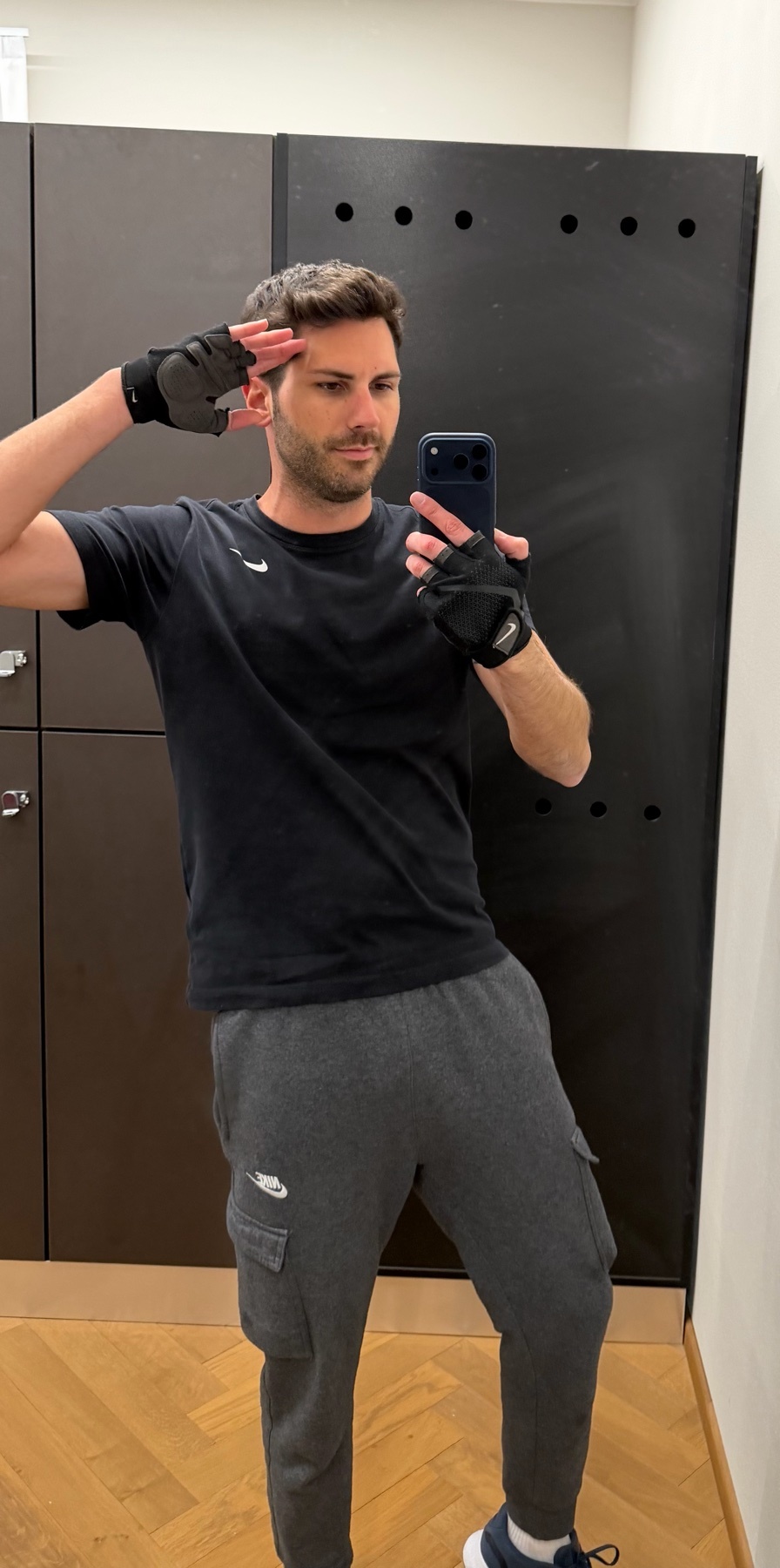 A man takes a selfie in a gym, wearing workout clothes. He is posing with his hand on his head.