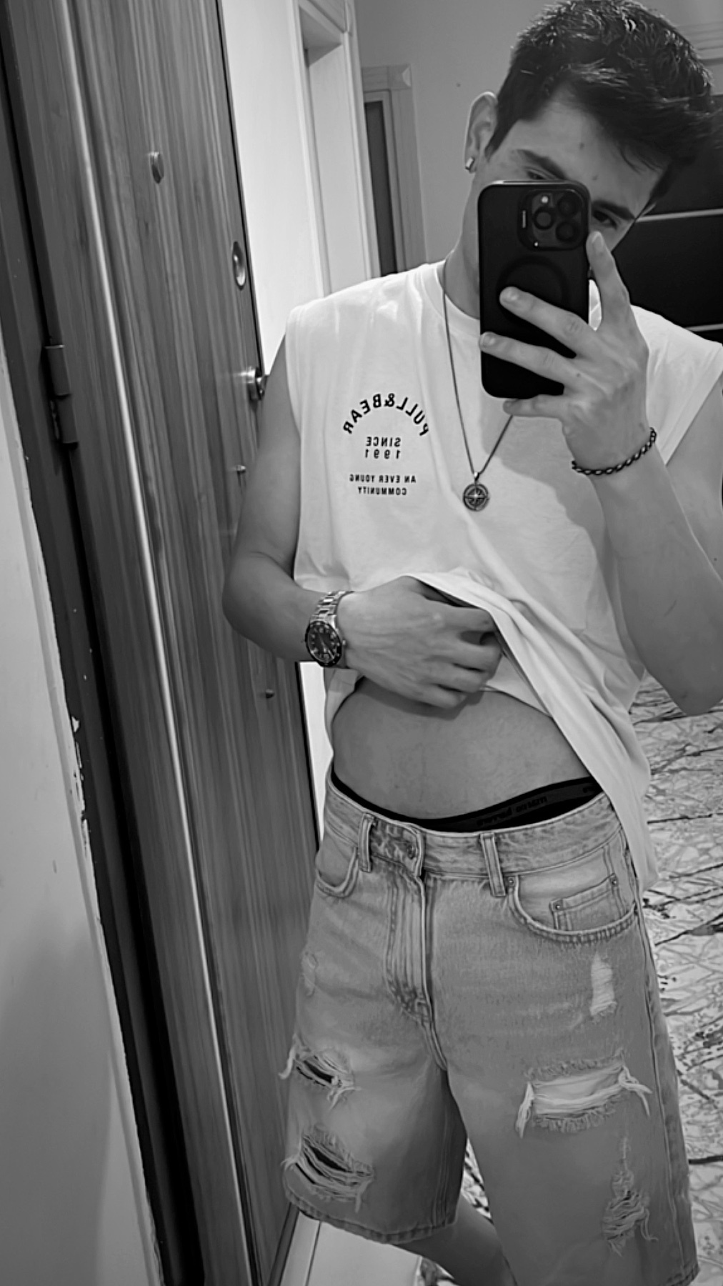 A man takes a mirror selfie, showcasing his physique. He's wearing a sleeveless t-shirt and ripped denim shorts.