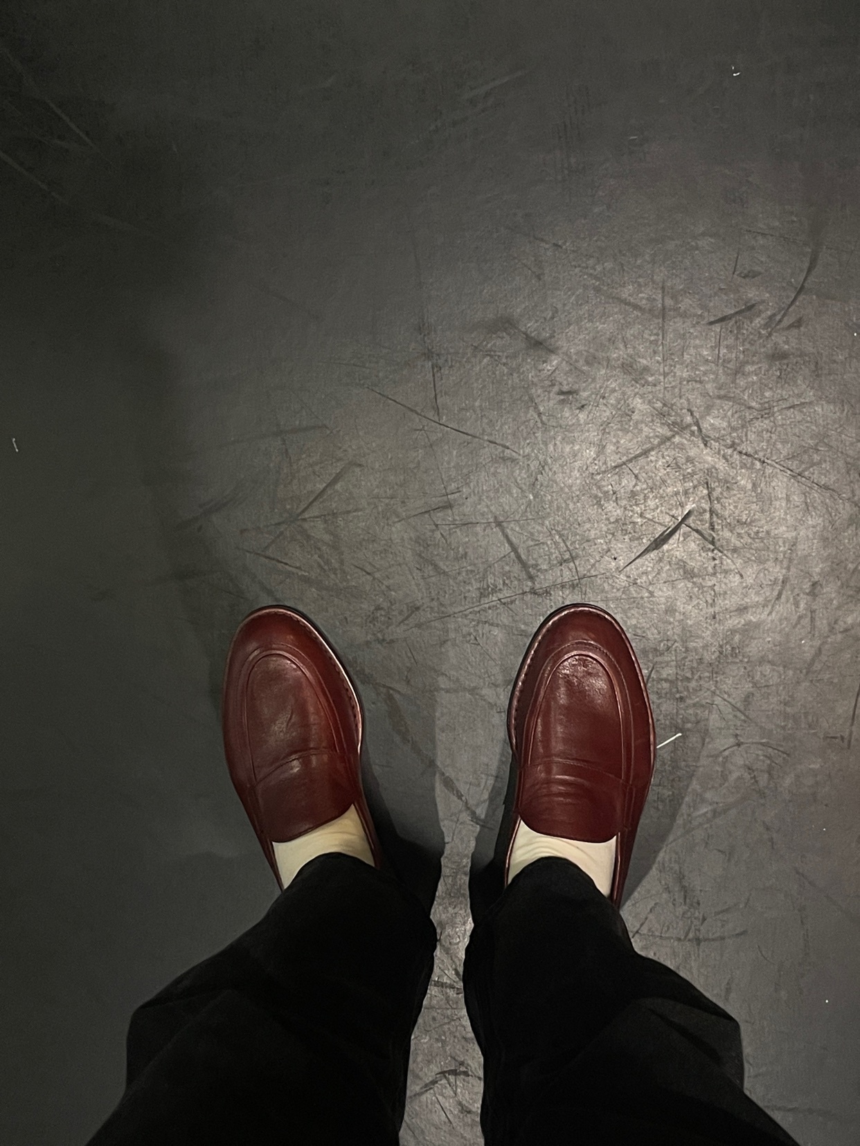 A top-down shot of a person's feet wearing red leather loafers and black pants. The shoes are the focal point against a dark, textured background.