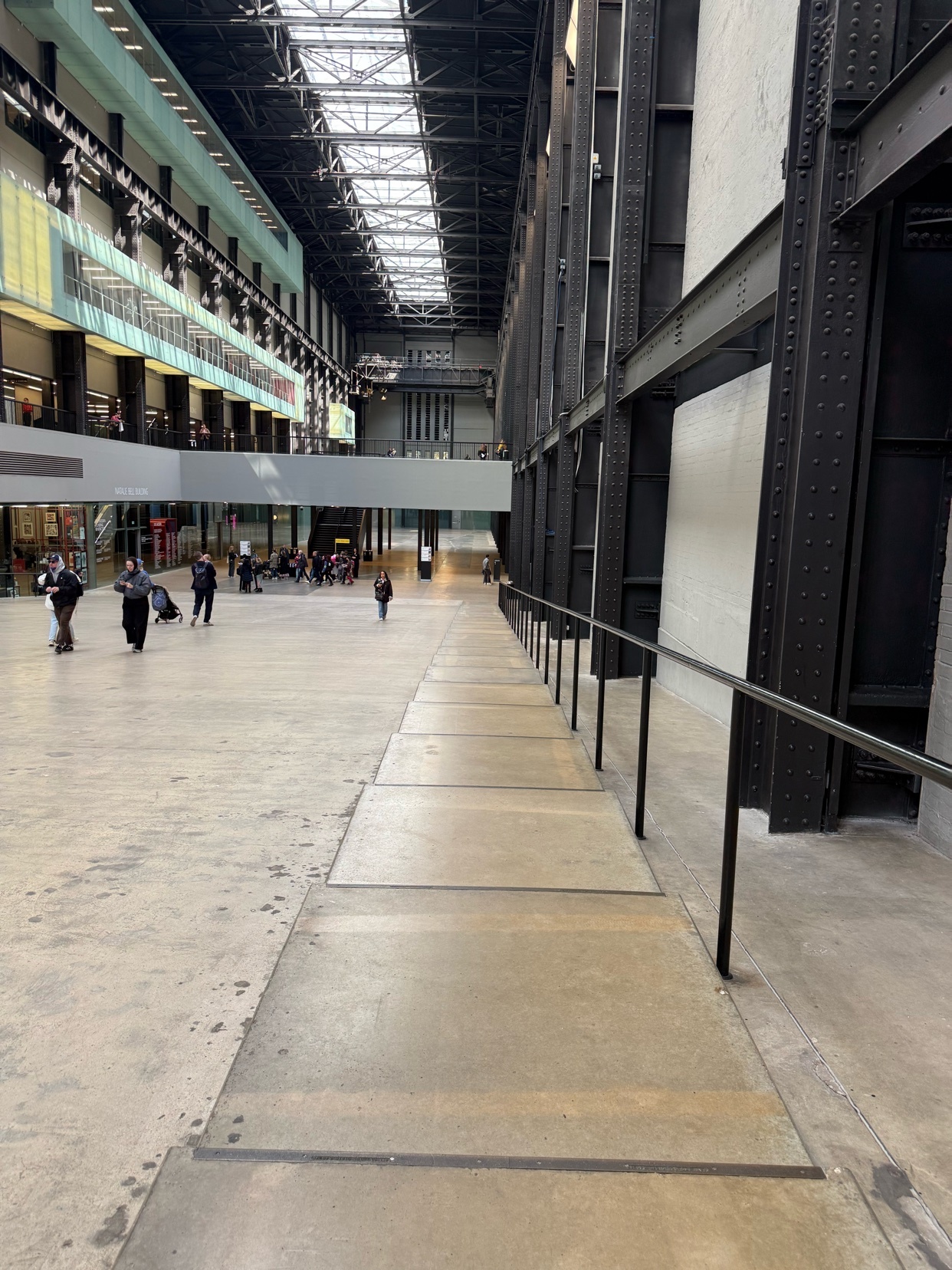 A wide, industrial-style interior space with high ceilings and exposed metal beams. People are seen walking in the distance, and natural light streams in from a large skylight.