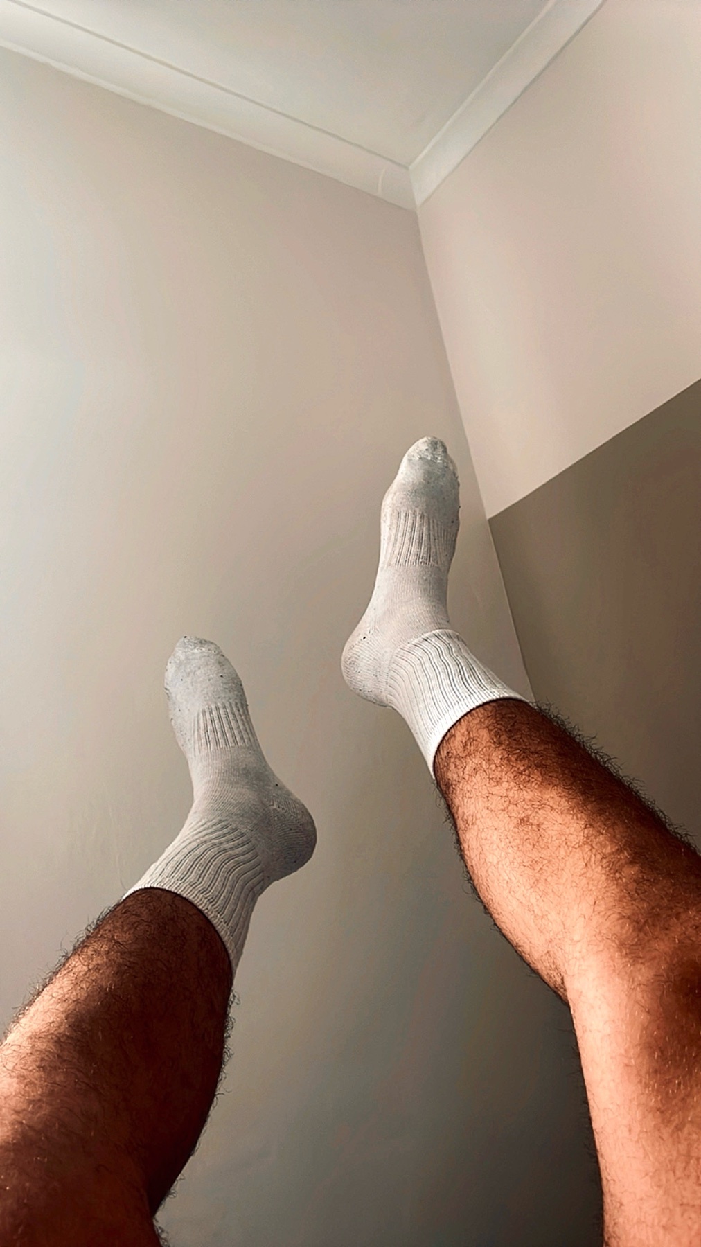 A low-angle shot shows a man's legs and feet wearing white socks against a neutral-colored wall.