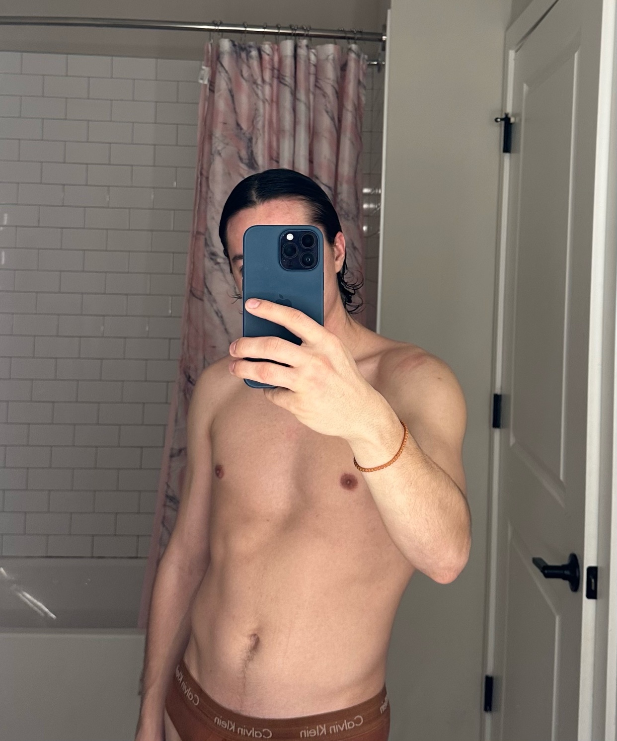 A man takes a shirtless mirror selfie in a bathroom. He is holding his phone up to take the photo.