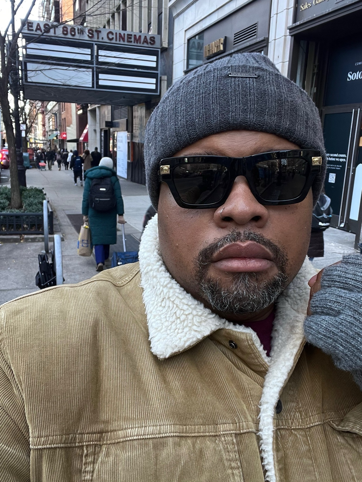 A man takes a selfie on a city street. He wears a beanie, sunglasses, and a corduroy jacket.