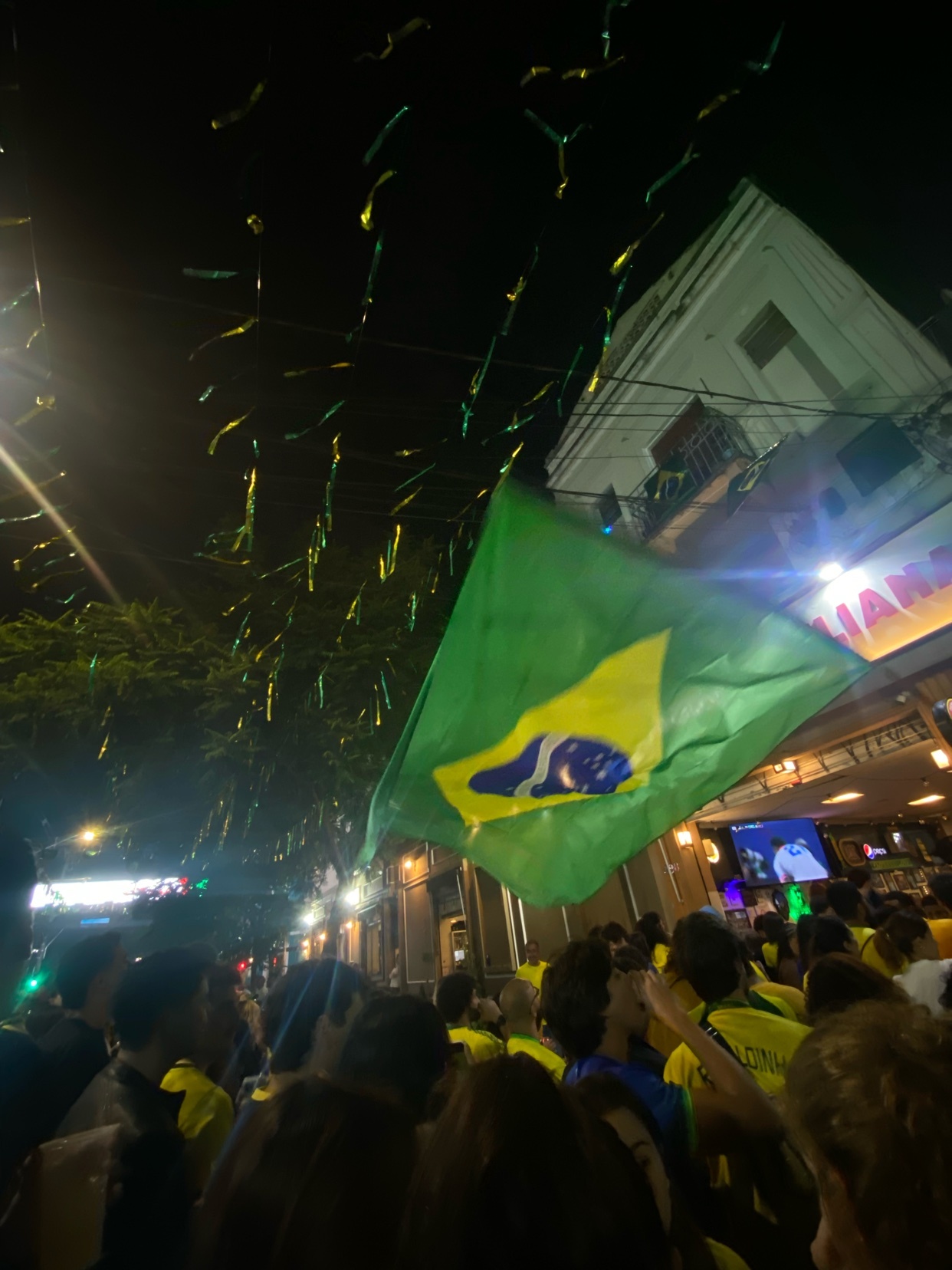 A Brazilian flag waves in the night sky above a crowded street lined with buildings and trees. Green and yellow streamers hang overhead, and people in yellow shirts are visible below.