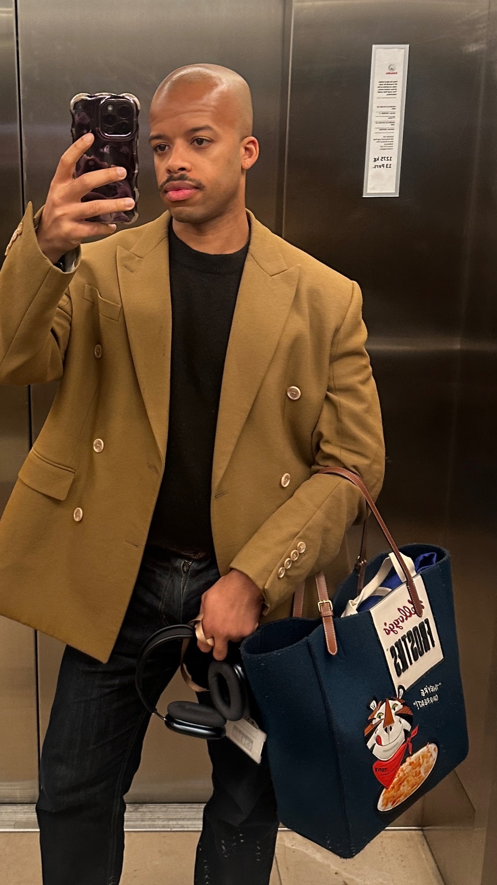 A man in a tan blazer and dark jeans takes a selfie in an elevator, holding a tote bag and headphones.
