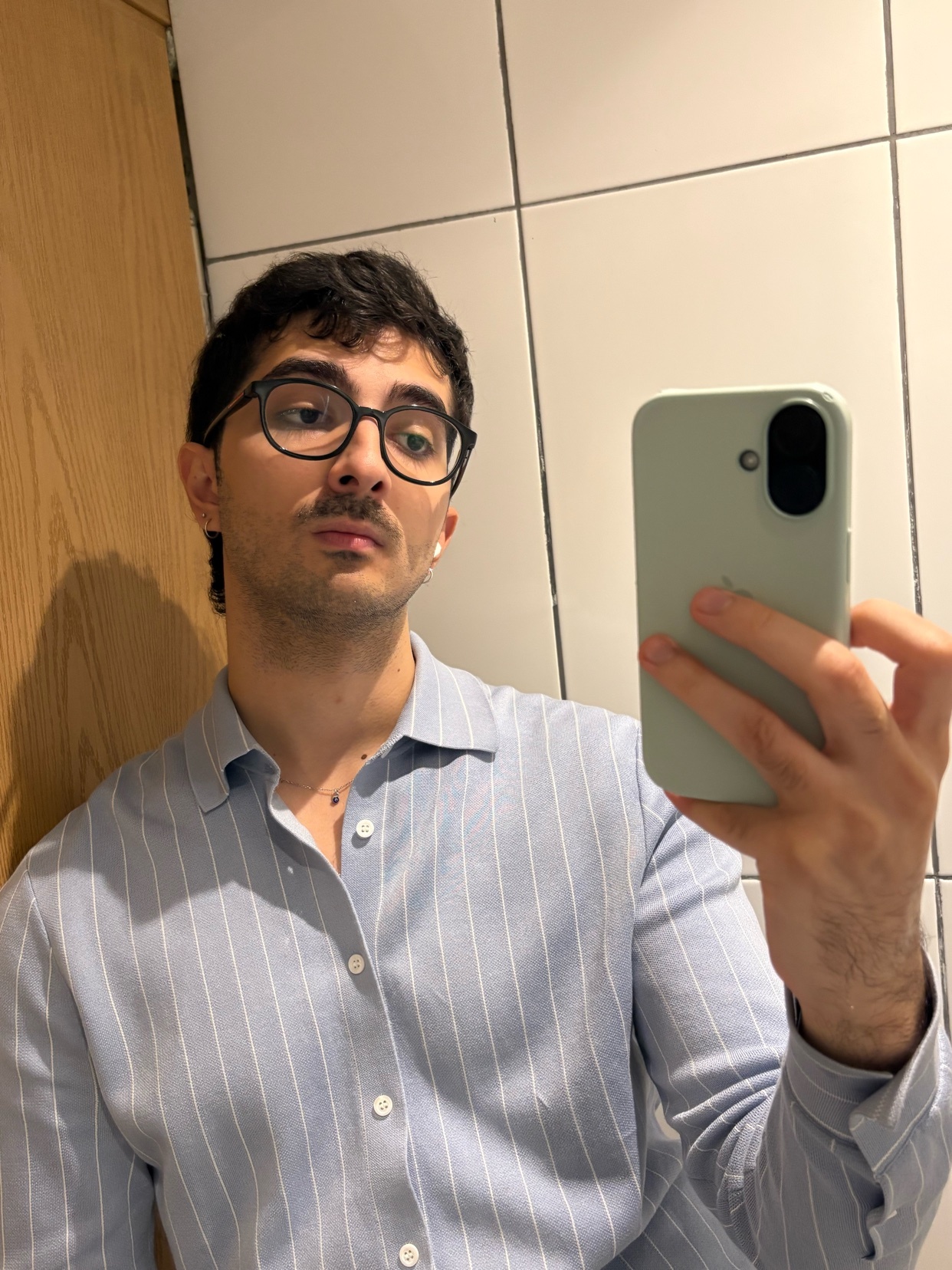 A man takes a selfie in a bathroom mirror. He is wearing glasses and a button-down shirt.