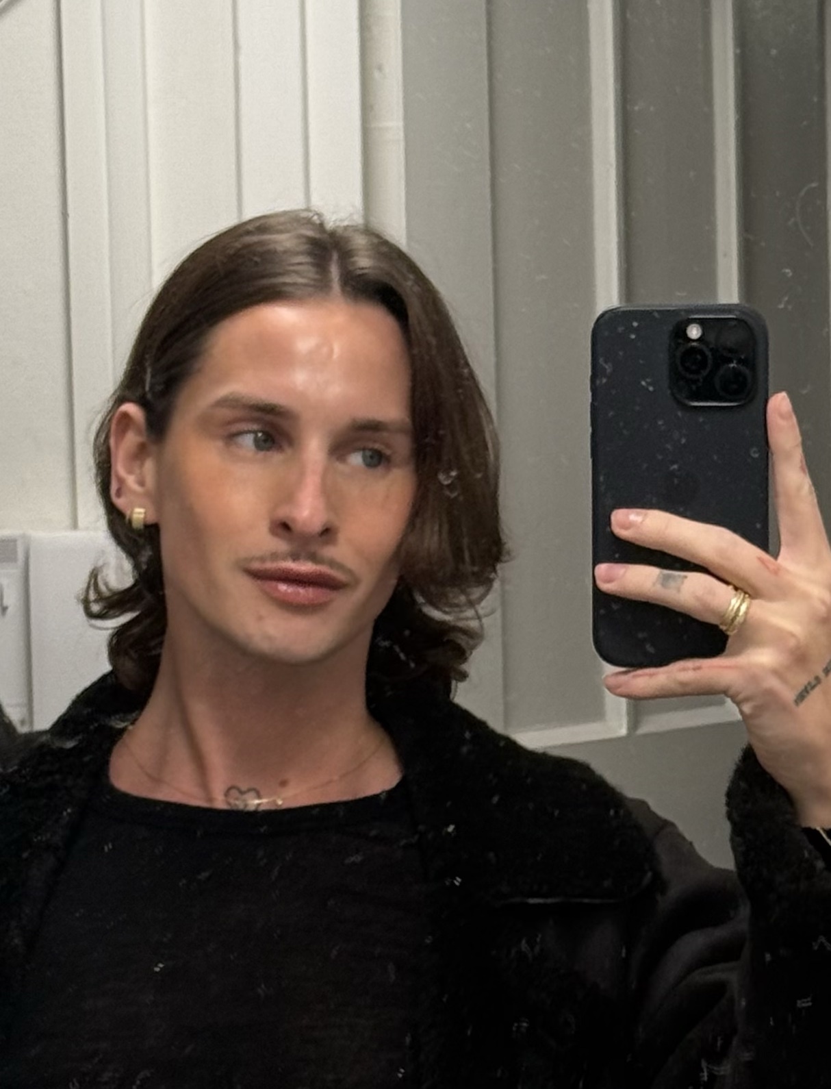 A man takes a selfie in a mirror. He has dark hair and a mustache, and is wearing jewelry.