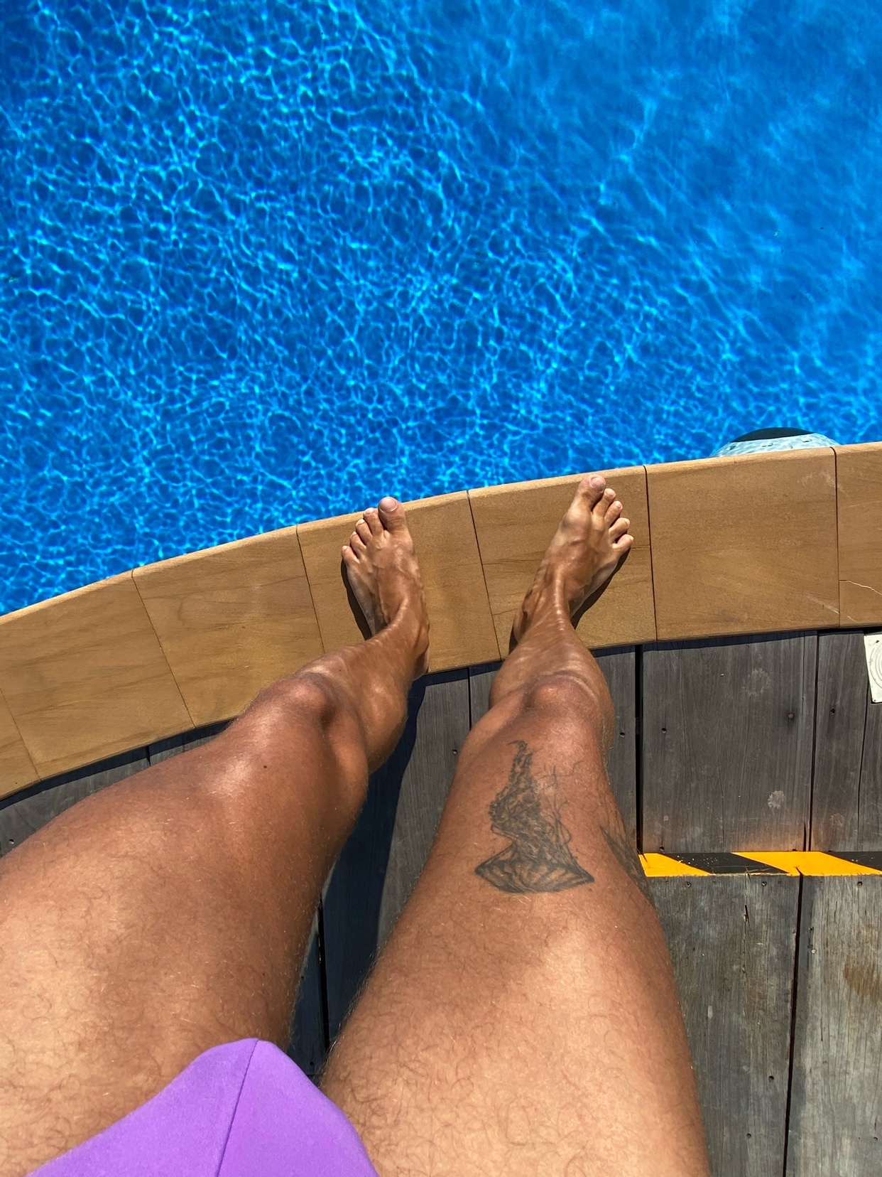 A man's legs are seen from a high angle, with a tattoo on his thigh. He is shirtless and wearing purple swimwear, with a pool in the background.