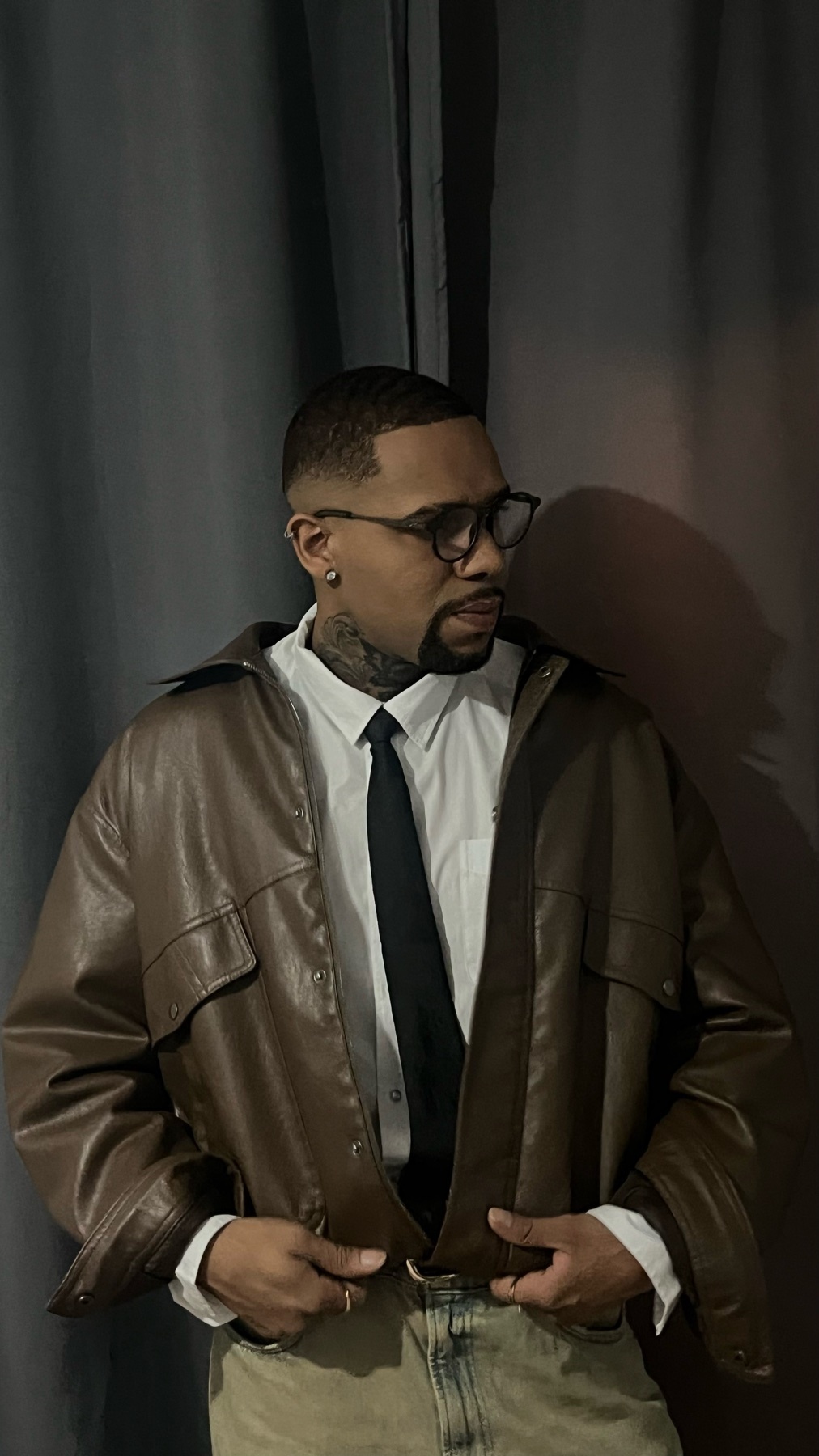 A man with tattoos on his neck and face poses in a brown leather jacket over a white shirt and tie. He wears glasses and has a beard.